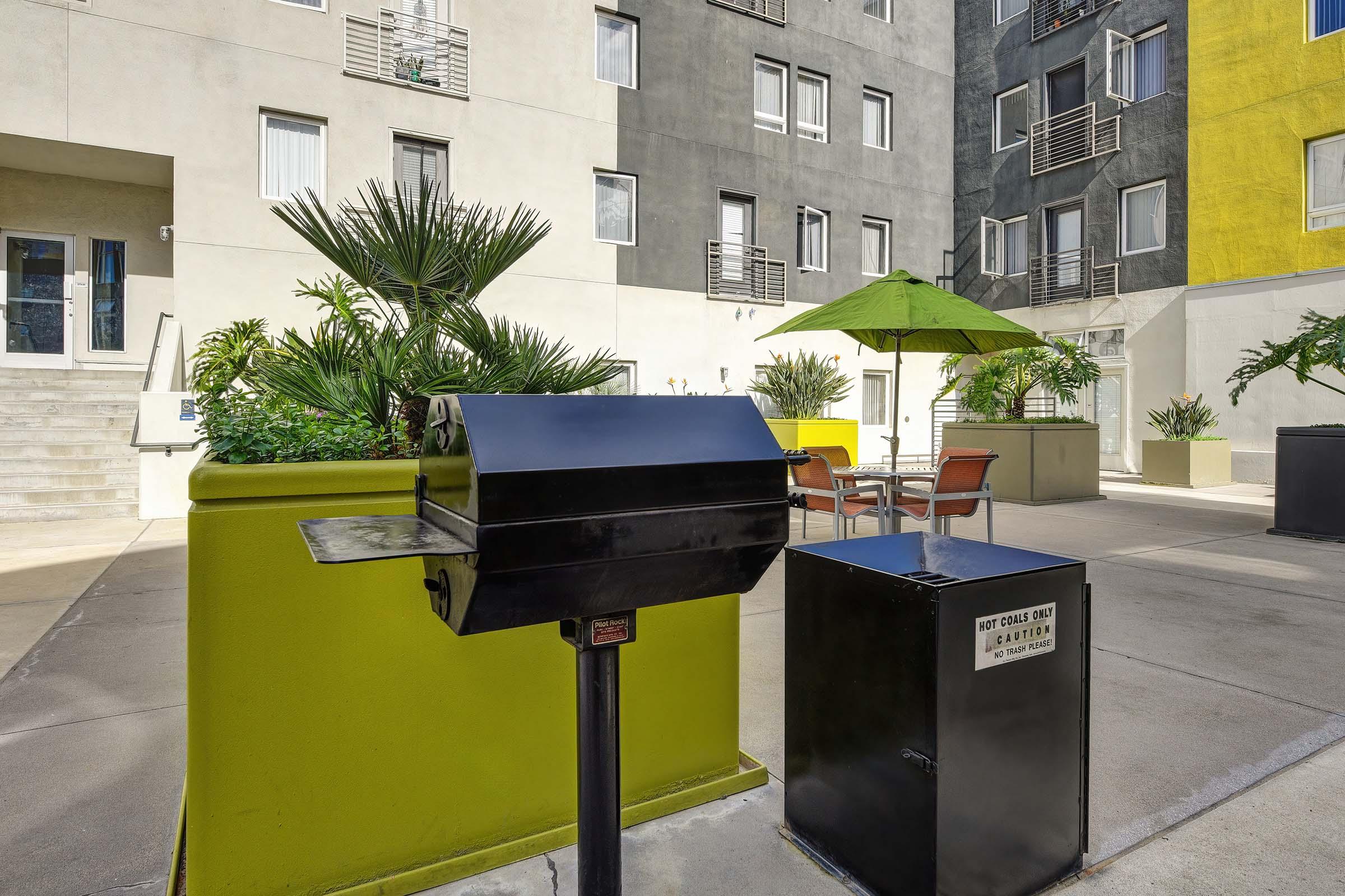 An outdoor courtyard featuring a black grill near vibrant green planters. In the background, there are apartment building walls in gray and yellow, with a patio set that includes an umbrella and chairs. Lush palm trees and other plants add greenery to the space.