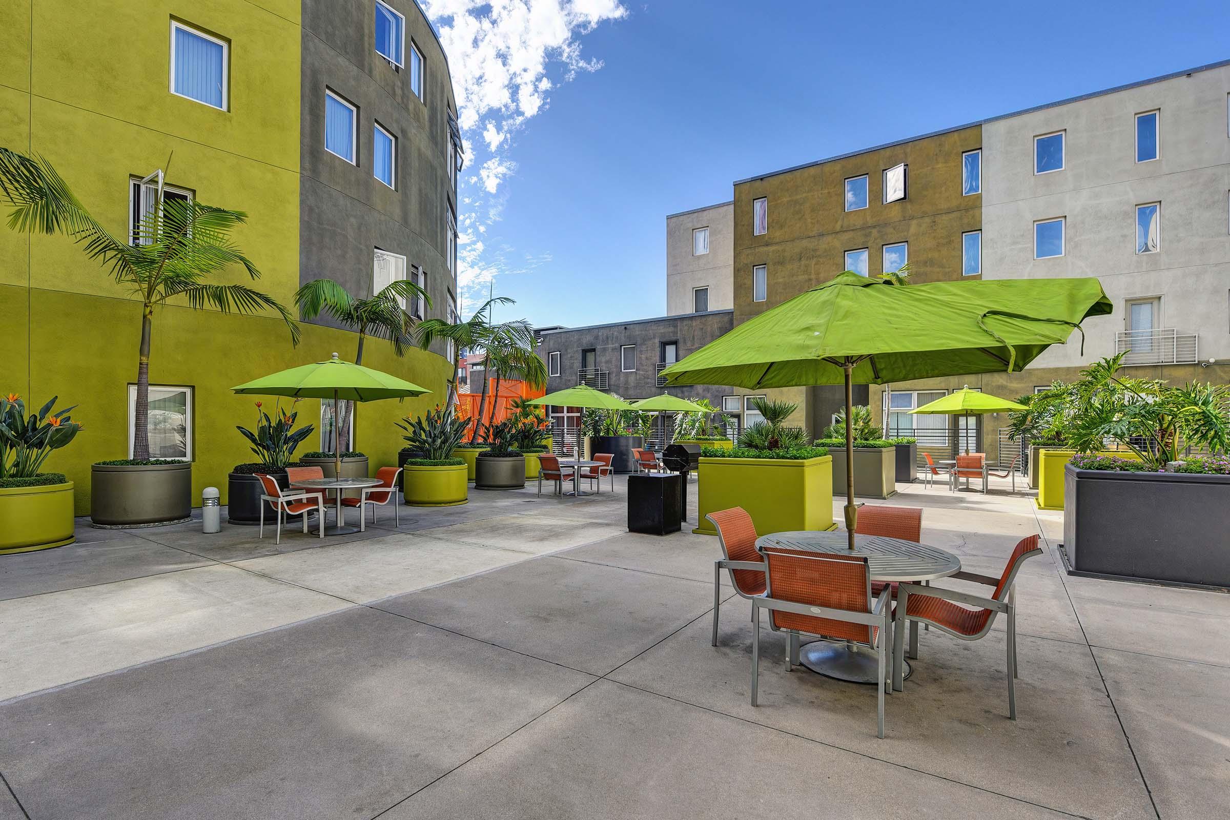 A vibrant outdoor courtyard featuring green umbrellas over patio tables surrounded by plants. The walls of the buildings are a mix of yellow and gray, and there are seating areas for relaxation. The sky is clear with a few clouds, creating a bright and inviting atmosphere.