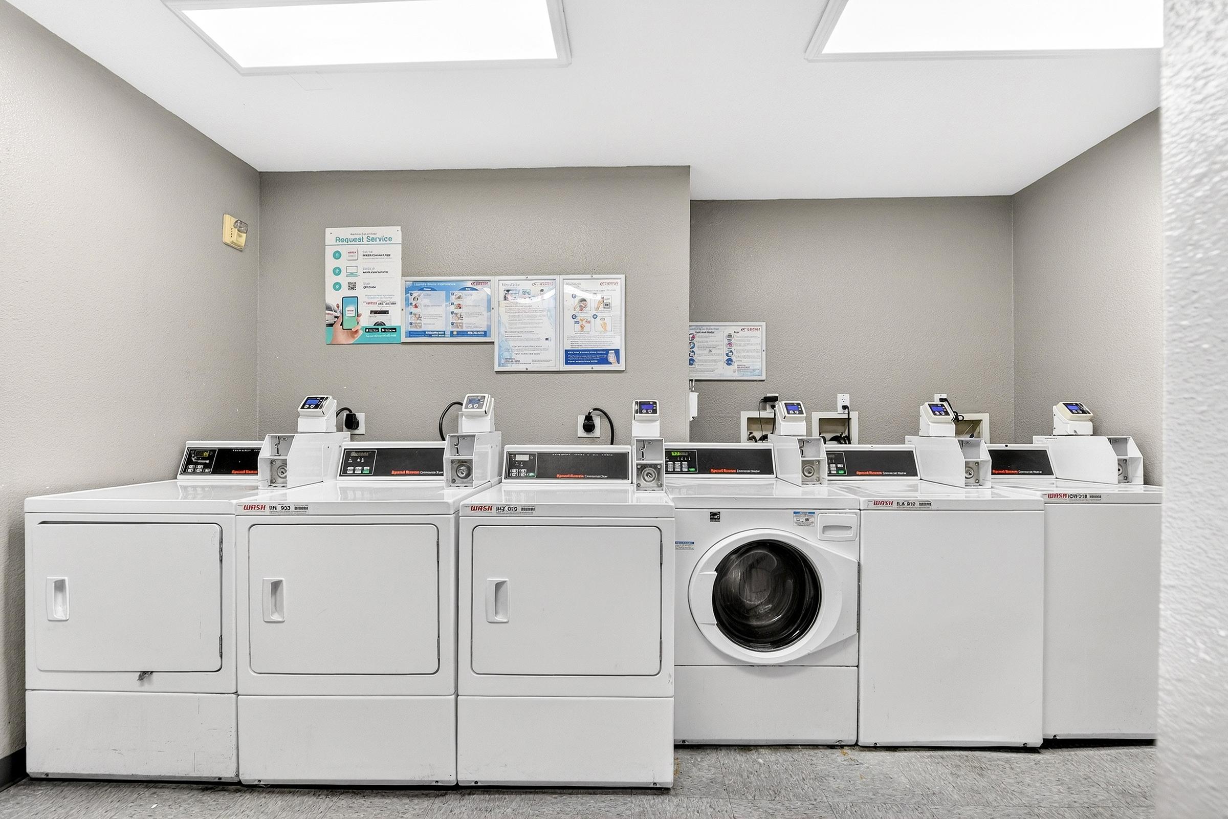 A clean and modern laundry room featuring multiple washing machines and dryers. The machines are lined up against a gray wall, with informative posters displayed nearby. Bright overhead lights illuminate the space, creating a welcoming atmosphere for laundry tasks.