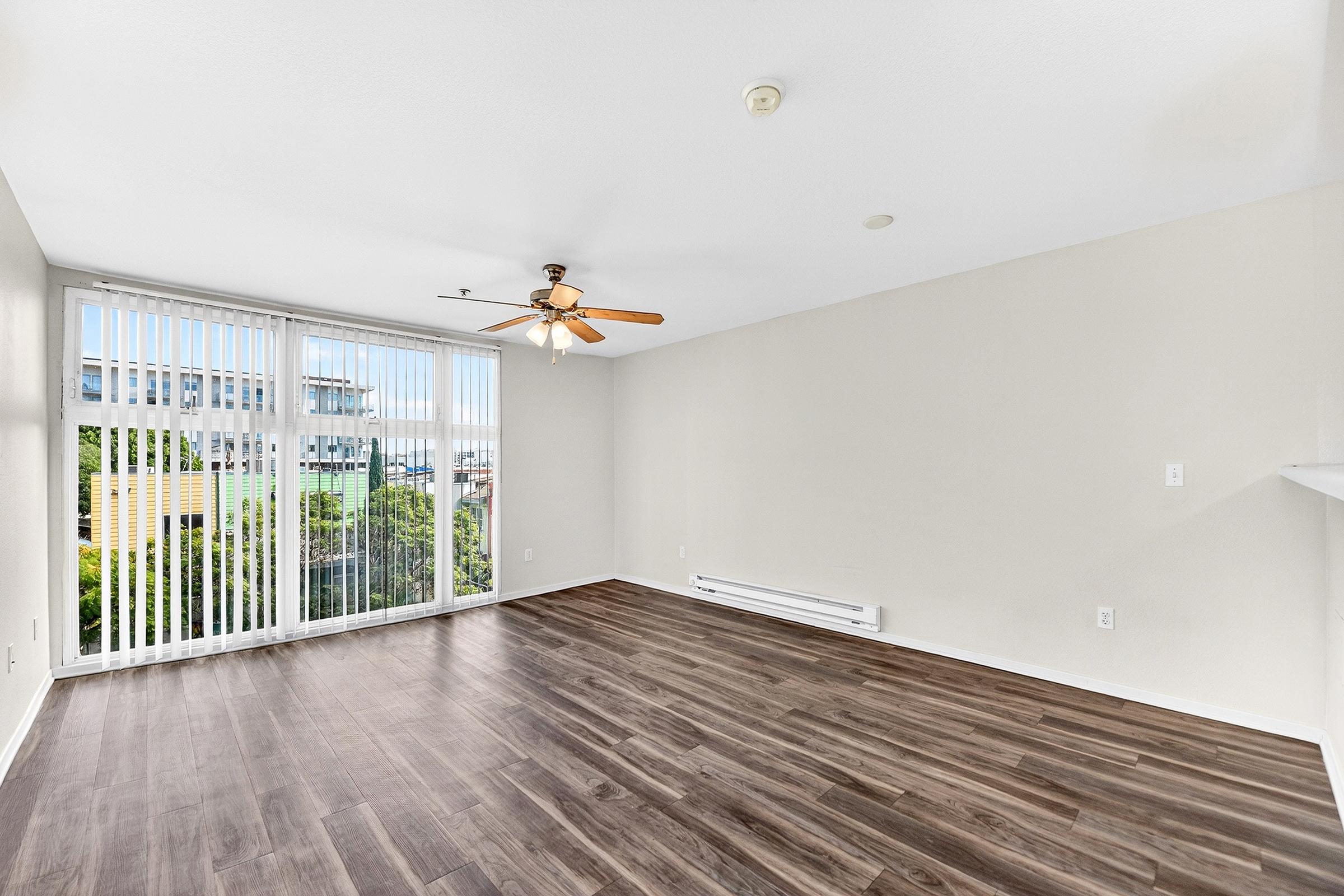 Bright and spacious living area featuring large windows with vertical blinds, a ceiling fan, and modern laminate flooring. The walls are painted a light neutral color, creating an inviting atmosphere. A heating unit is visible along the wall, and views of greenery can be seen through the windows.