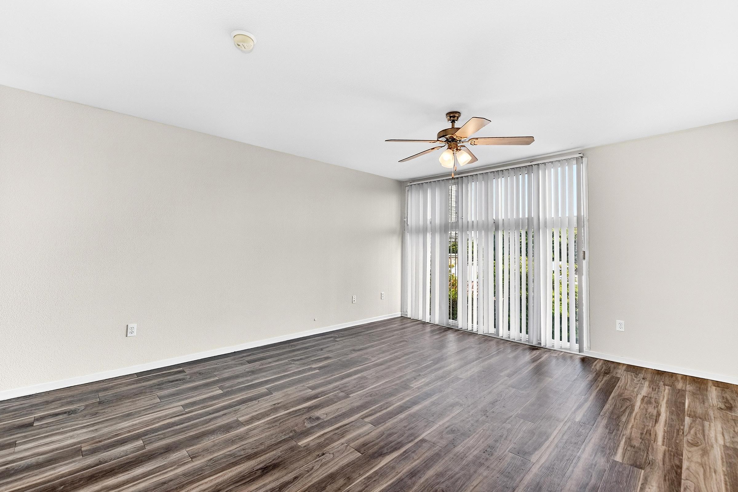 Empty living room with light-colored walls and dark wood laminate flooring. A ceiling fan is visible, and large windows with vertical blinds allow natural light to enter the space. The room appears spacious and unfurnished, creating an inviting atmosphere for personalization.