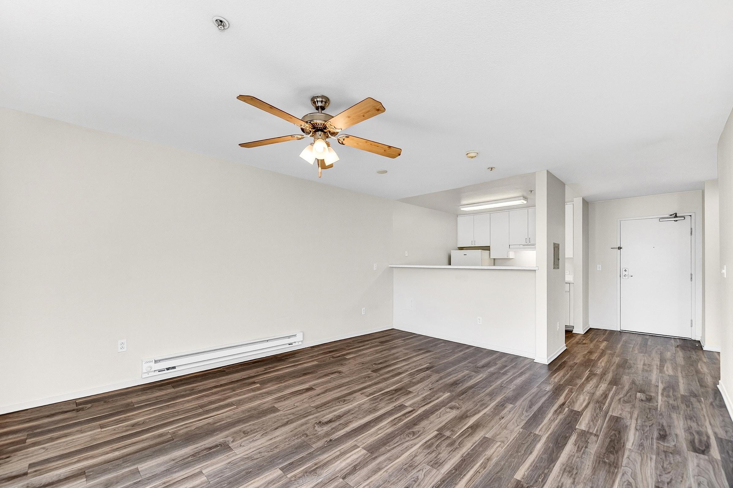 Spacious living area featuring a ceiling fan and hardwood-style flooring. The layout includes a kitchenette with light-colored cabinets and appliances. A door leading outside is visible, along with a baseboard heater. The walls are painted in a neutral tone, creating a bright and open atmosphere.
