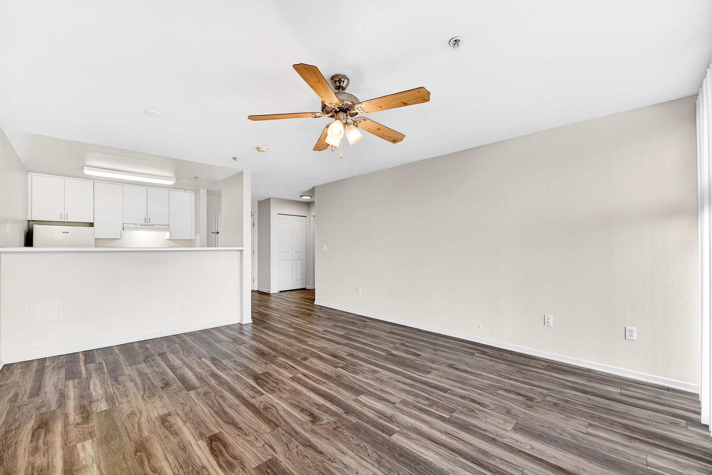 Spacious and light-filled apartment interior featuring a living area with a ceiling fan, hardwood-style flooring, and light-colored walls. The kitchen area is visible in the background, showcasing modern cabinetry and appliances. The open layout enhances the sense of space and invites natural light.