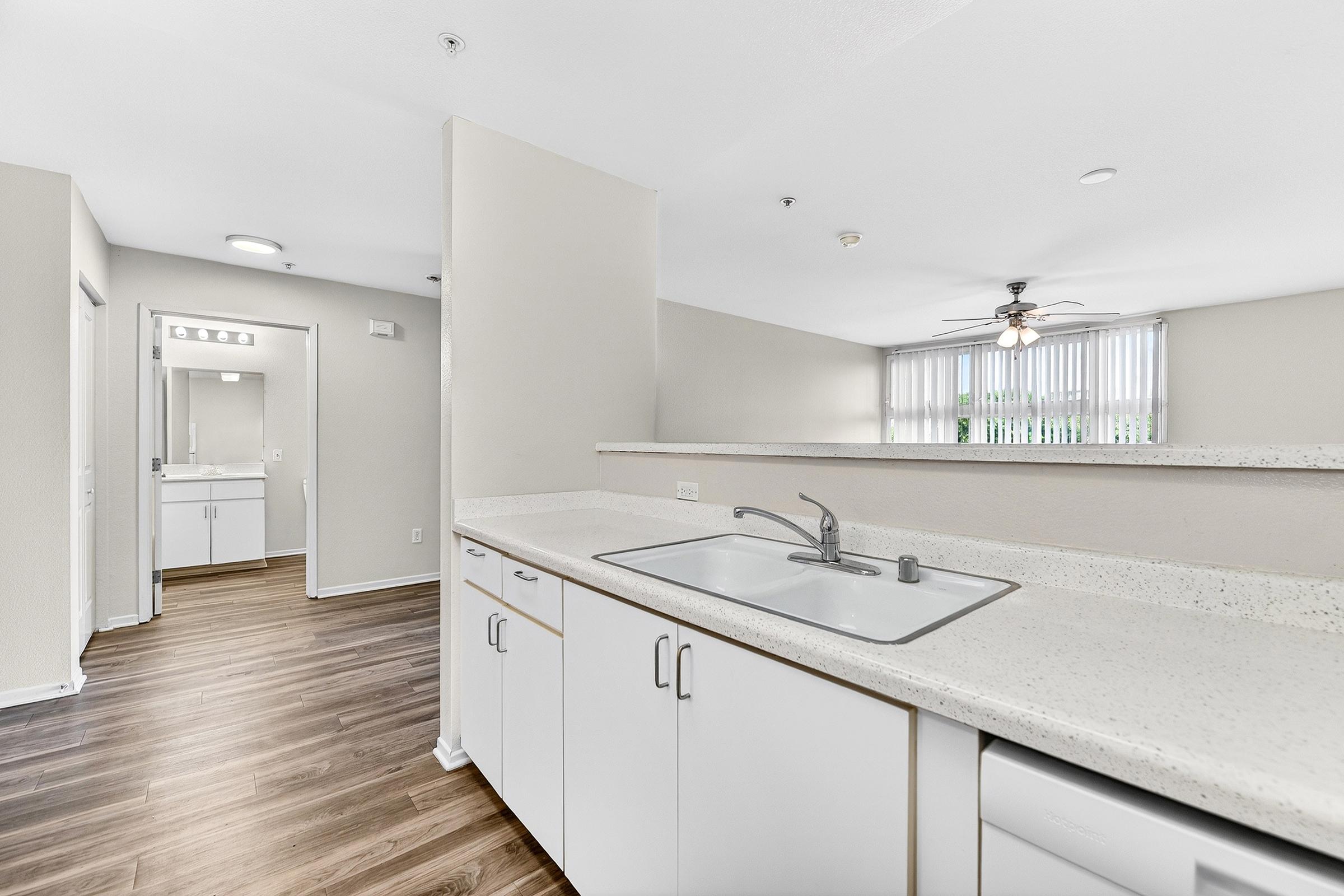 A modern kitchen with white cabinets, a stainless steel sink, and a countertop overlooking an open living space. Light wood flooring extends into the adjacent area, which features natural light from windows. A ceiling fan is visible, and a bathroom can be seen in the background through an open door.