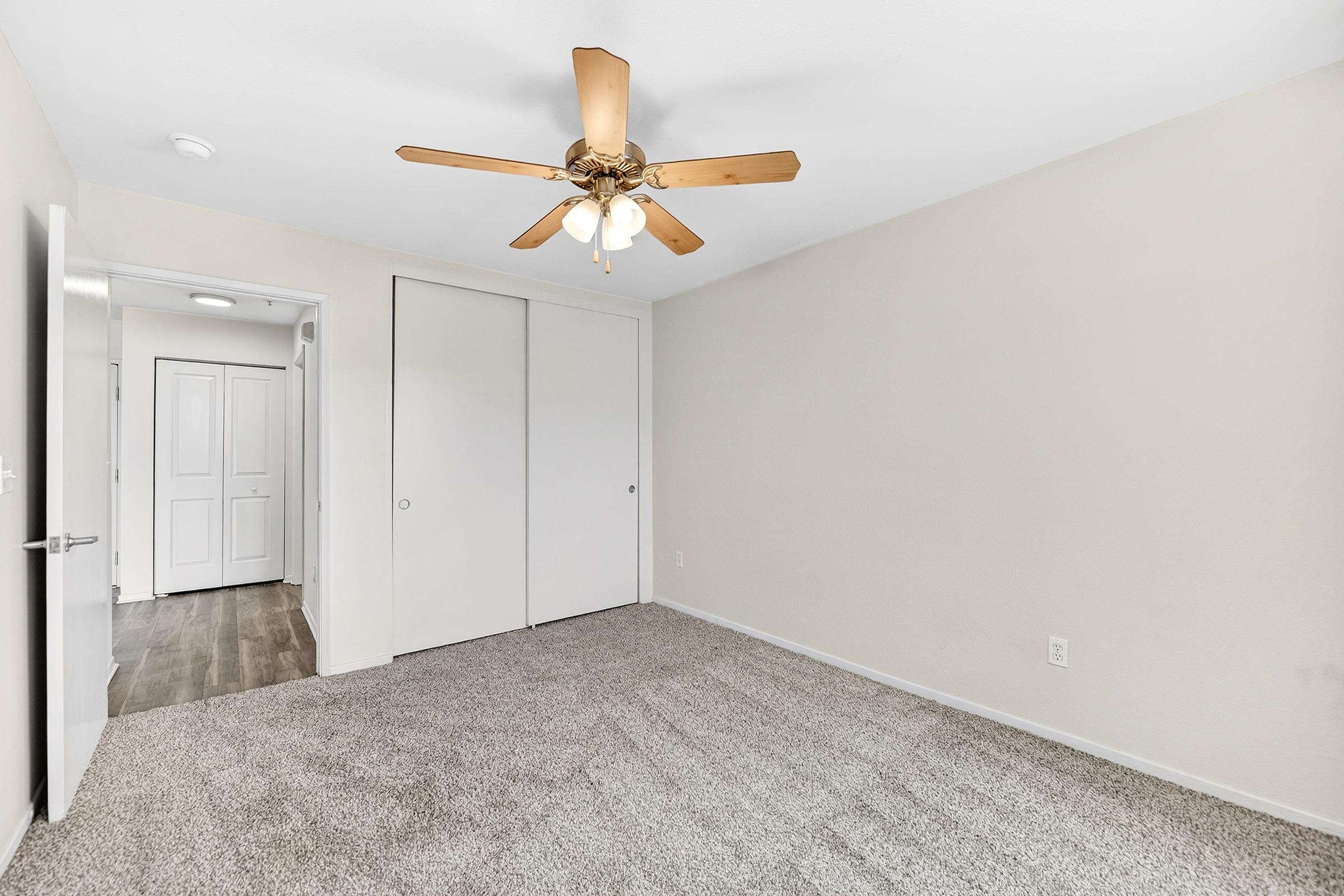 Empty room with light beige walls and plush carpet. A ceiling fan with wooden blades hangs in the center. There's a closed closet with sliding doors on one side, and a white door leading to another room is visible in the background. Natural light brightens the space, creating a neutral and airy atmosphere.