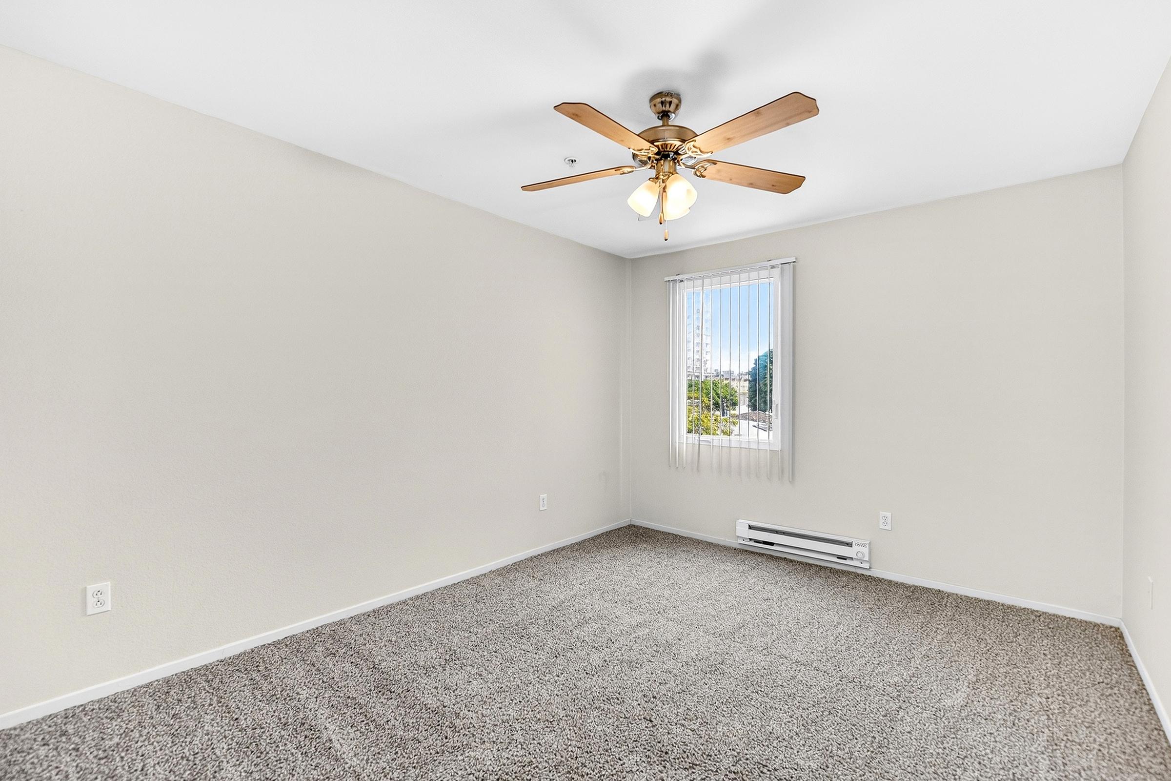 A spacious, empty room featuring light beige walls, a ceiling fan with wooden blades, and large windows with vertical blinds. The floor is covered with a soft, tan carpet. There is a heating unit along one wall, and the overall atmosphere is bright and airy.