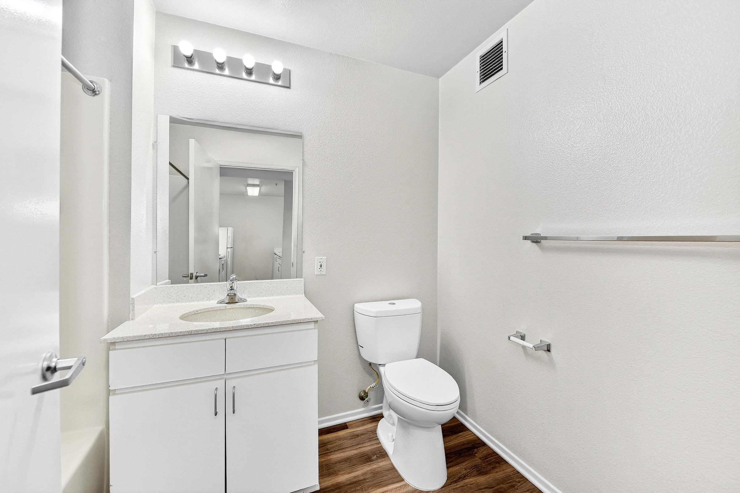 A modern, clean bathroom featuring a white toilet and a sink with a granite countertop. The space has light-colored walls, a large mirror above the sink, and a simple lighting fixture with three bulbs. There’s a towel bar and a small window in the background, enhancing the bright, airy feel of the room.