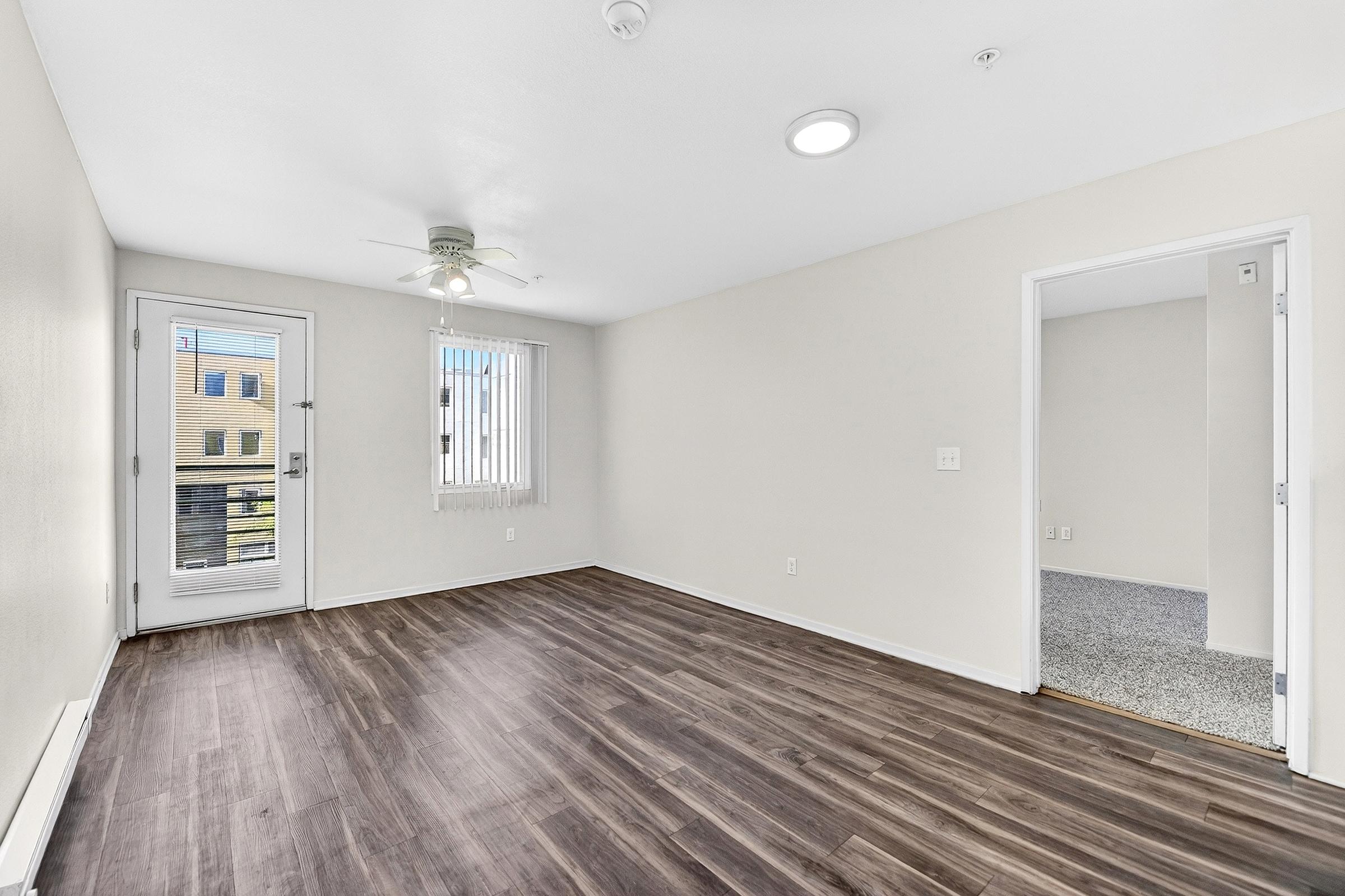 A spacious, empty living area with light wood flooring and neutral walls. A ceiling fan is mounted in the corner, and a door leads to a small balcony, while large windows allow natural light. A second room is visible in the background with carpeted flooring. The space is well-lit and ready for furnishings.