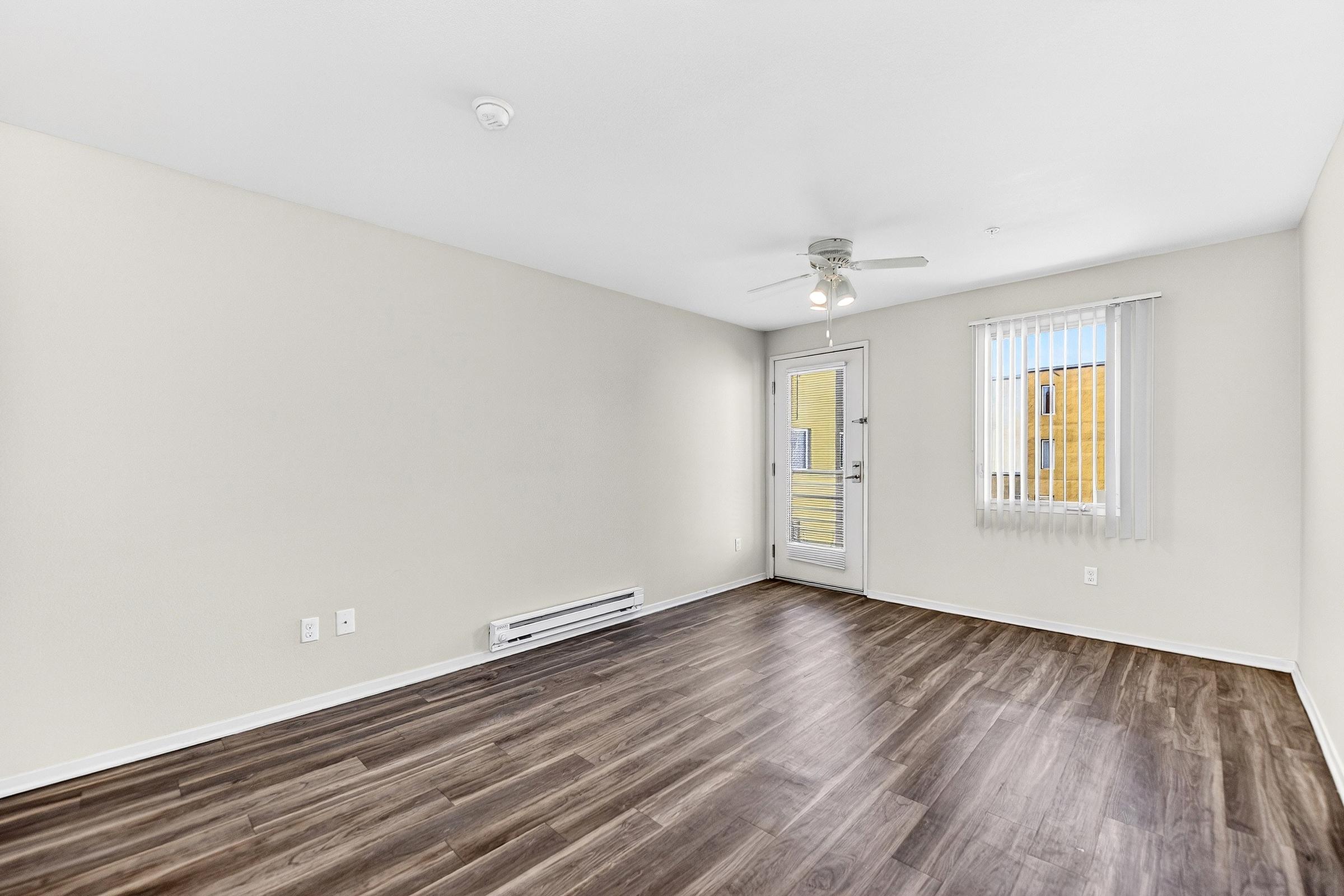 A spacious, unfurnished room featuring light-colored walls and dark wooden flooring. A ceiling fan is mounted on the ceiling, and a set of sliding glass doors leads to a small balcony. Vertical blinds cover a window, allowing natural light to enter the space.