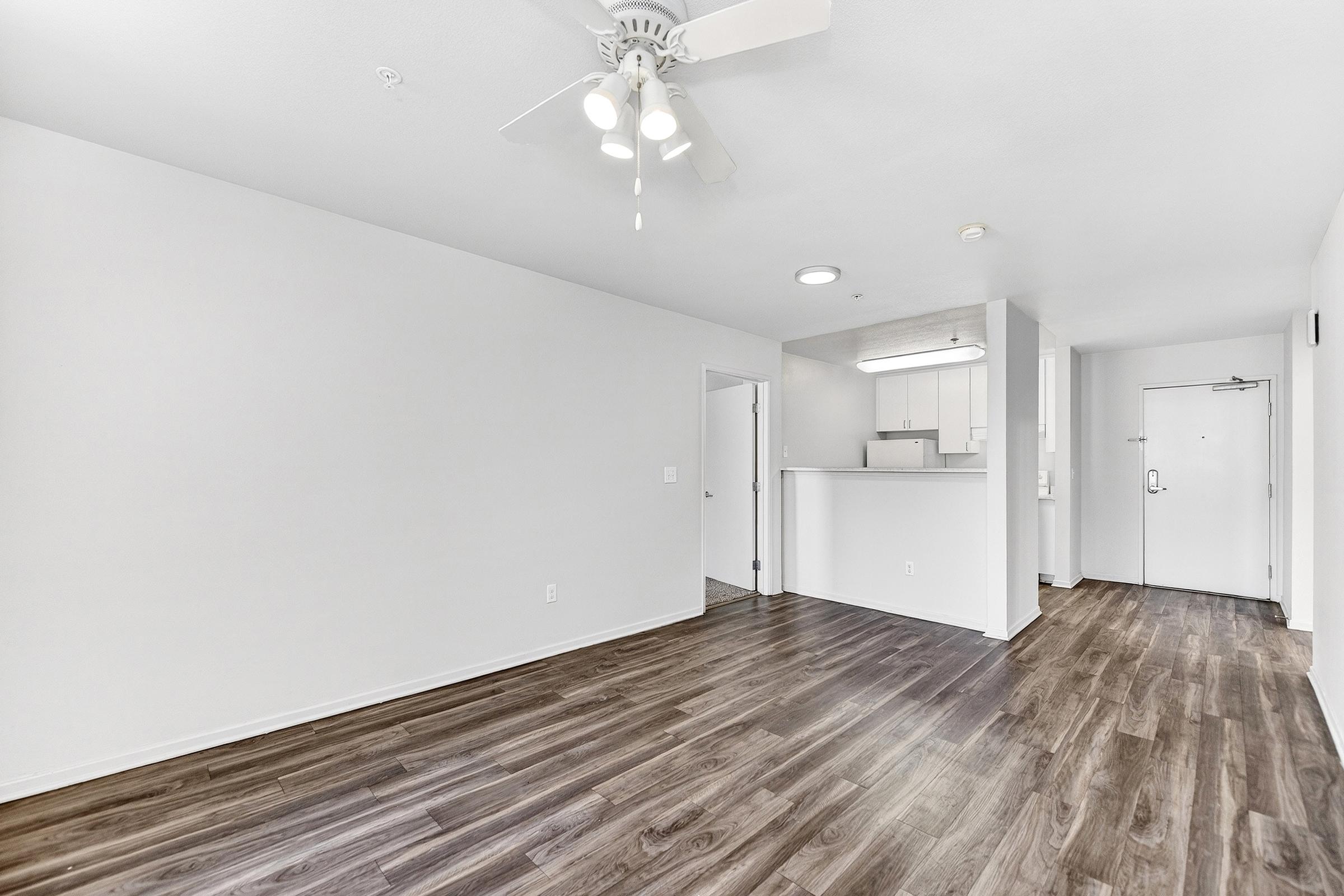 A spacious, light-filled living area with laminate flooring, featuring a ceiling fan. There is a doorway leading to the kitchen with bar seating, and another doorway leading to an exit. The walls are painted white, creating a clean and modern look.