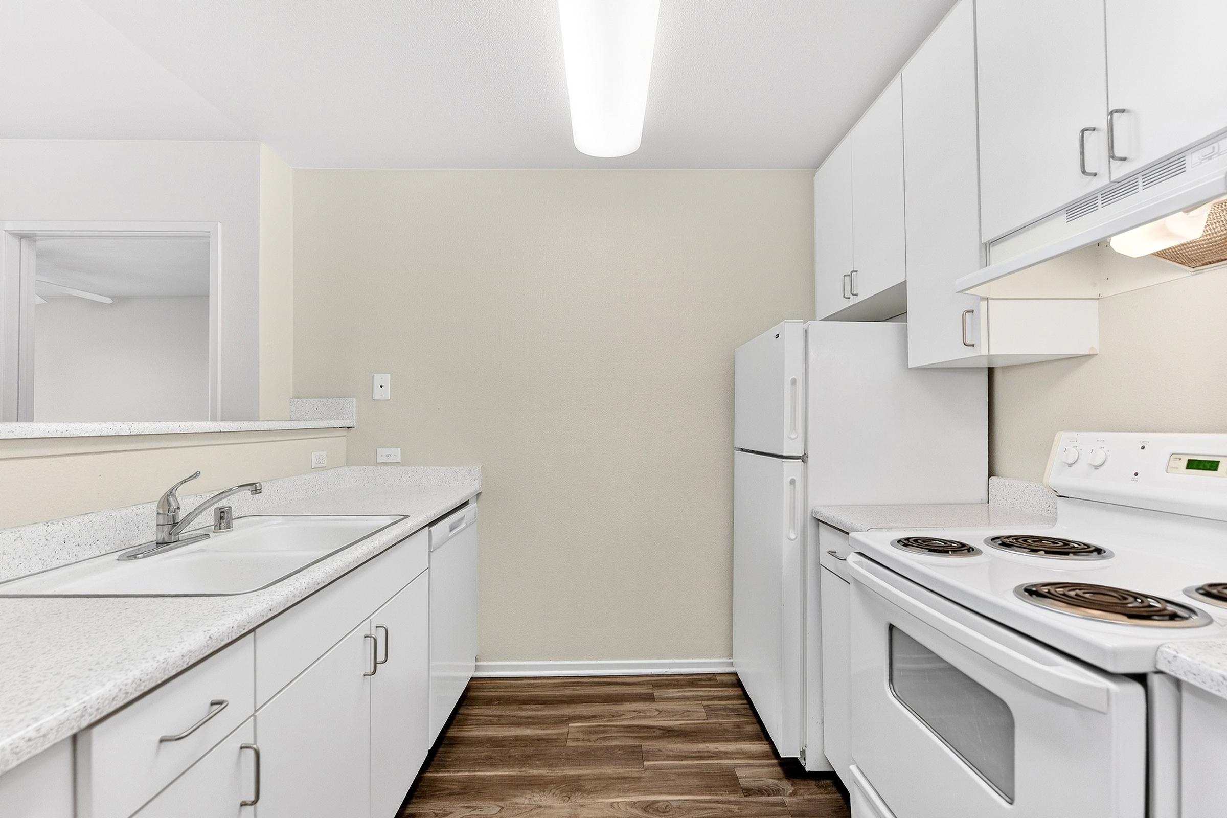 A modern kitchen featuring white cabinets, a double sink, and a white stove with four burners. The refrigerator stands to the right of the stove, and light wood flooring complements the overall bright and clean aesthetic of the space.