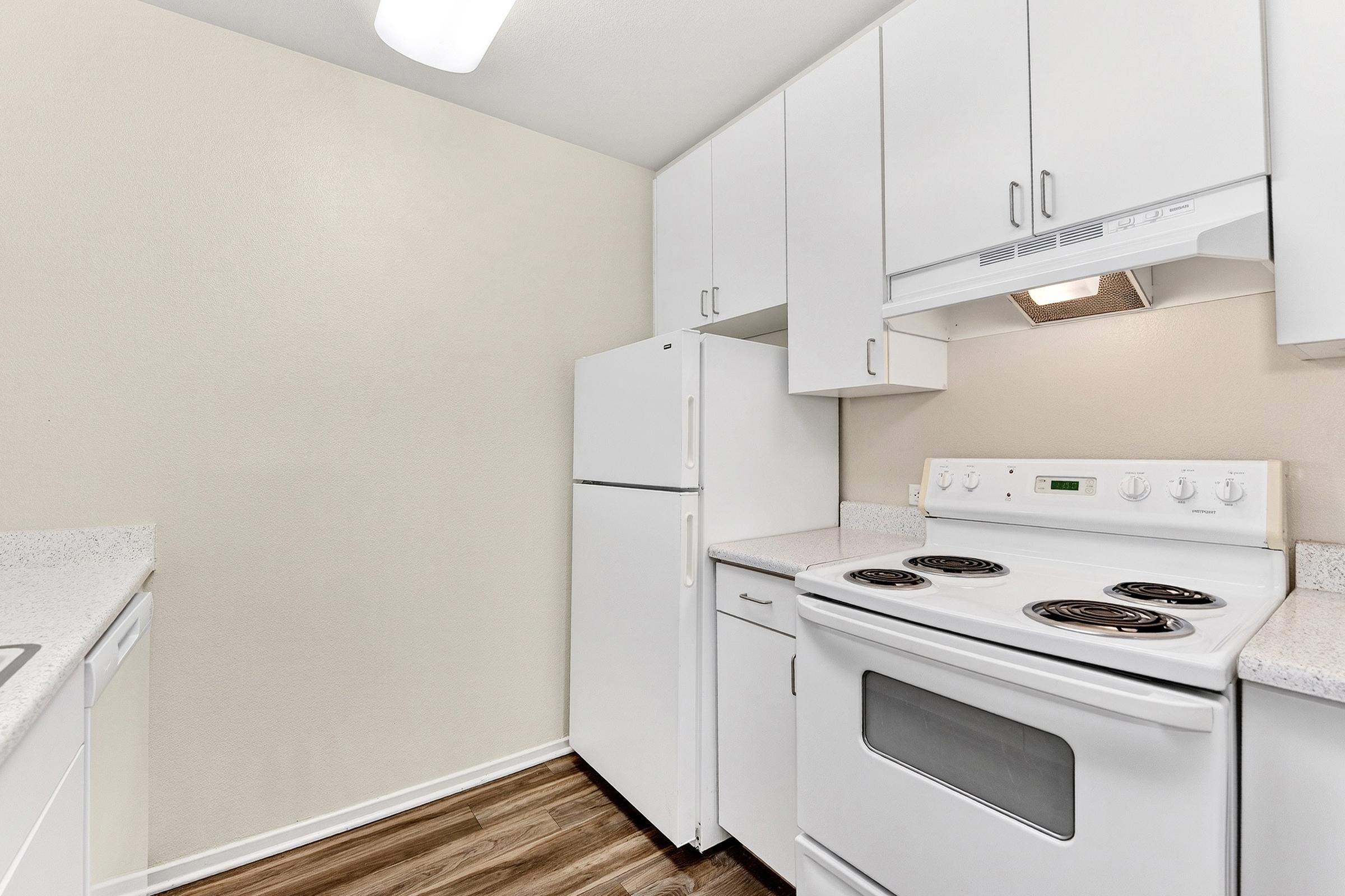 A clean, modern kitchen featuring white cabinets, a white refrigerator, and an electric stove. The countertops are light-colored, and the flooring is dark wood. The walls are painted a neutral tone, creating a bright and airy atmosphere. 