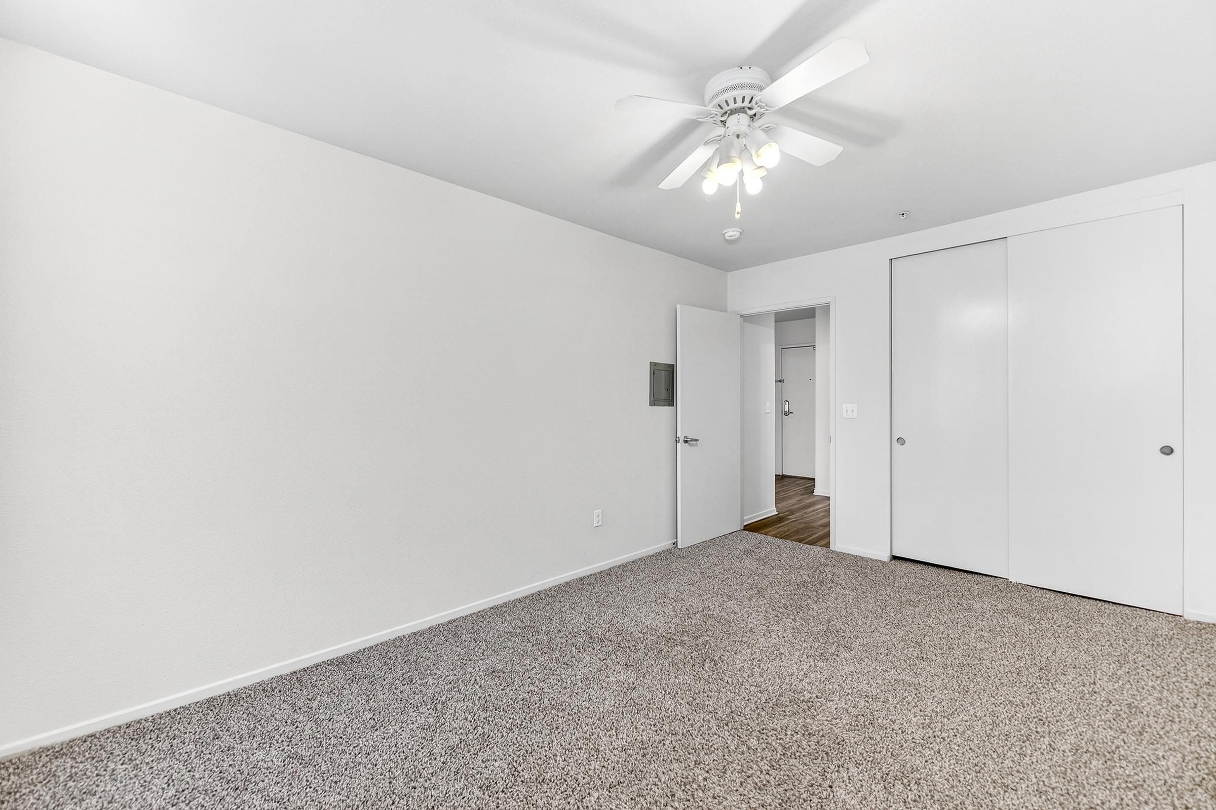 A spacious room with light beige carpet and white walls. There's a ceiling fan with lights, a closed closet with sliding doors on the right, and an entrance leading to another part of the space. The overall atmosphere is bright and inviting, suitable for various uses.