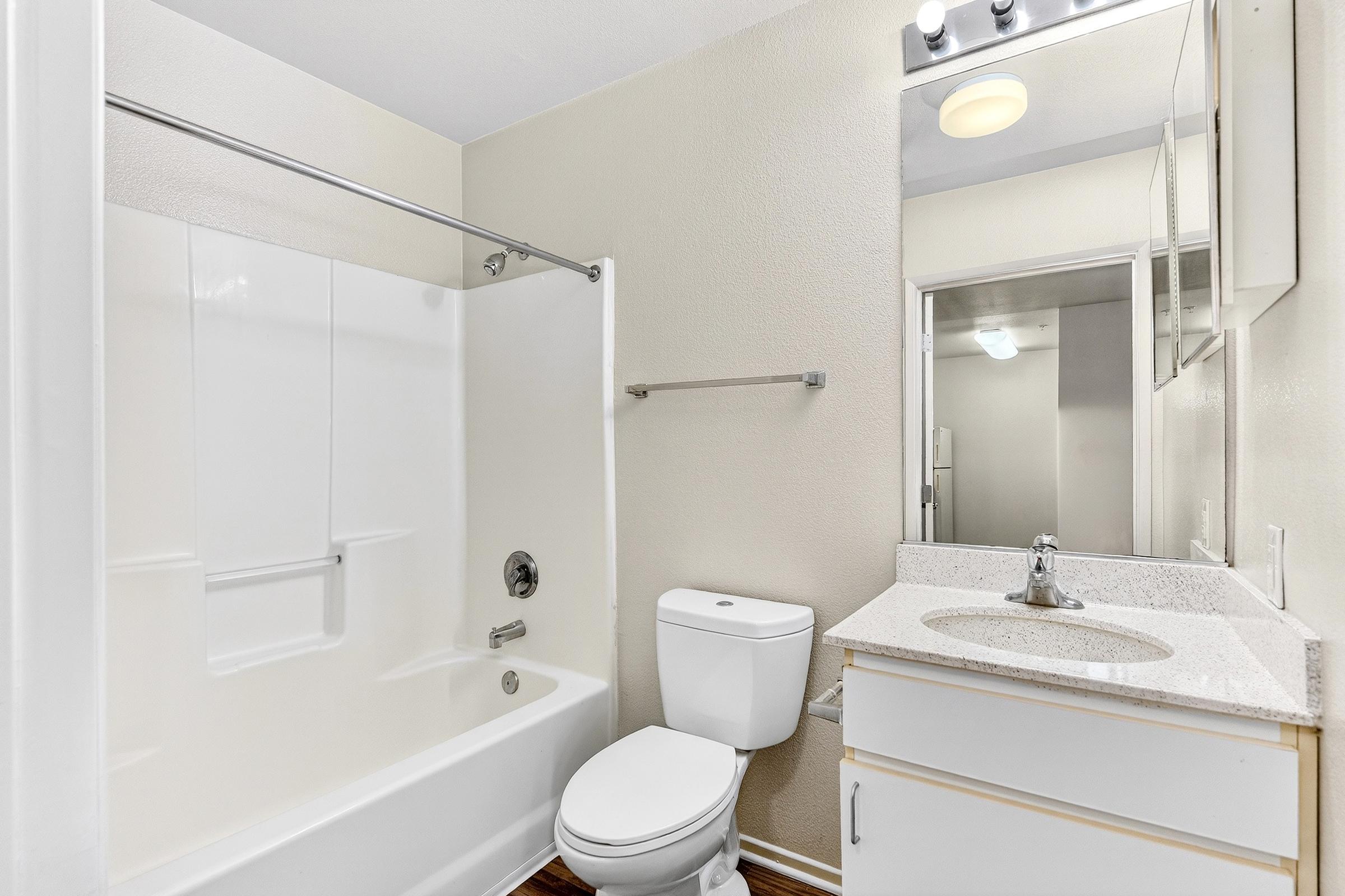 A clean, modern bathroom featuring a white bathtub with a shower, a toilet, and a sink with a light-colored countertop. The walls are painted in a soft beige, and a mirror reflects the space. Natural light is present, enhancing the bright and airy feel of the room.