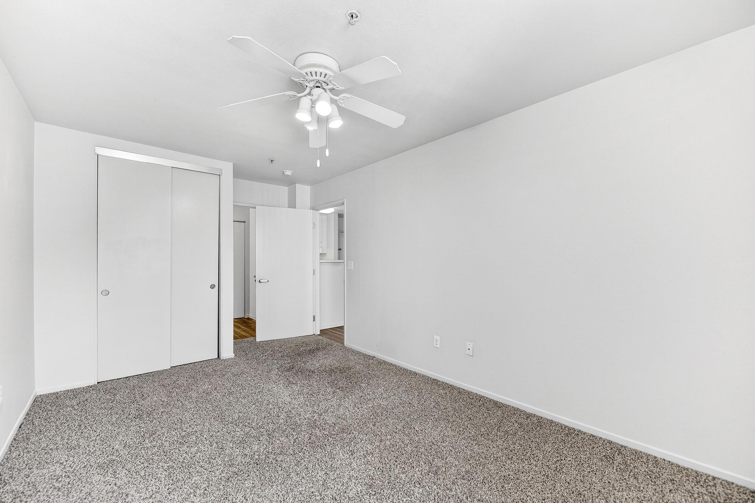 A spacious, unfurnished room with light gray walls and carpet flooring. It features a ceiling fan, a double closet with sliding doors, and an open doorway leading to another room. Natural light fills the space, giving it a bright and airy feel, ideal for decorating or use as a living area or bedroom.