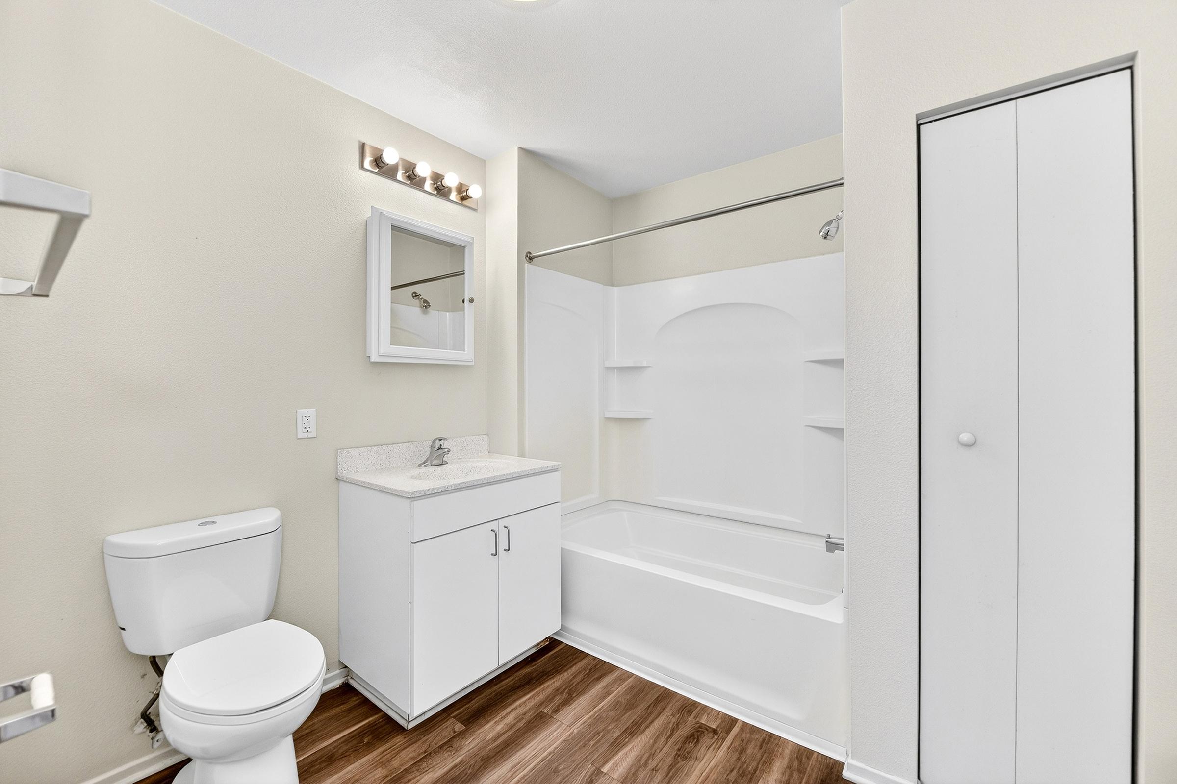 A clean, modern bathroom featuring a white toilet, a vanity with a sink and storage, a bathtub with a shower curtain, and a wall-mounted mirror. The walls are painted in a light color, and the floor is made of wood laminate. A small closet is visible on the right side.