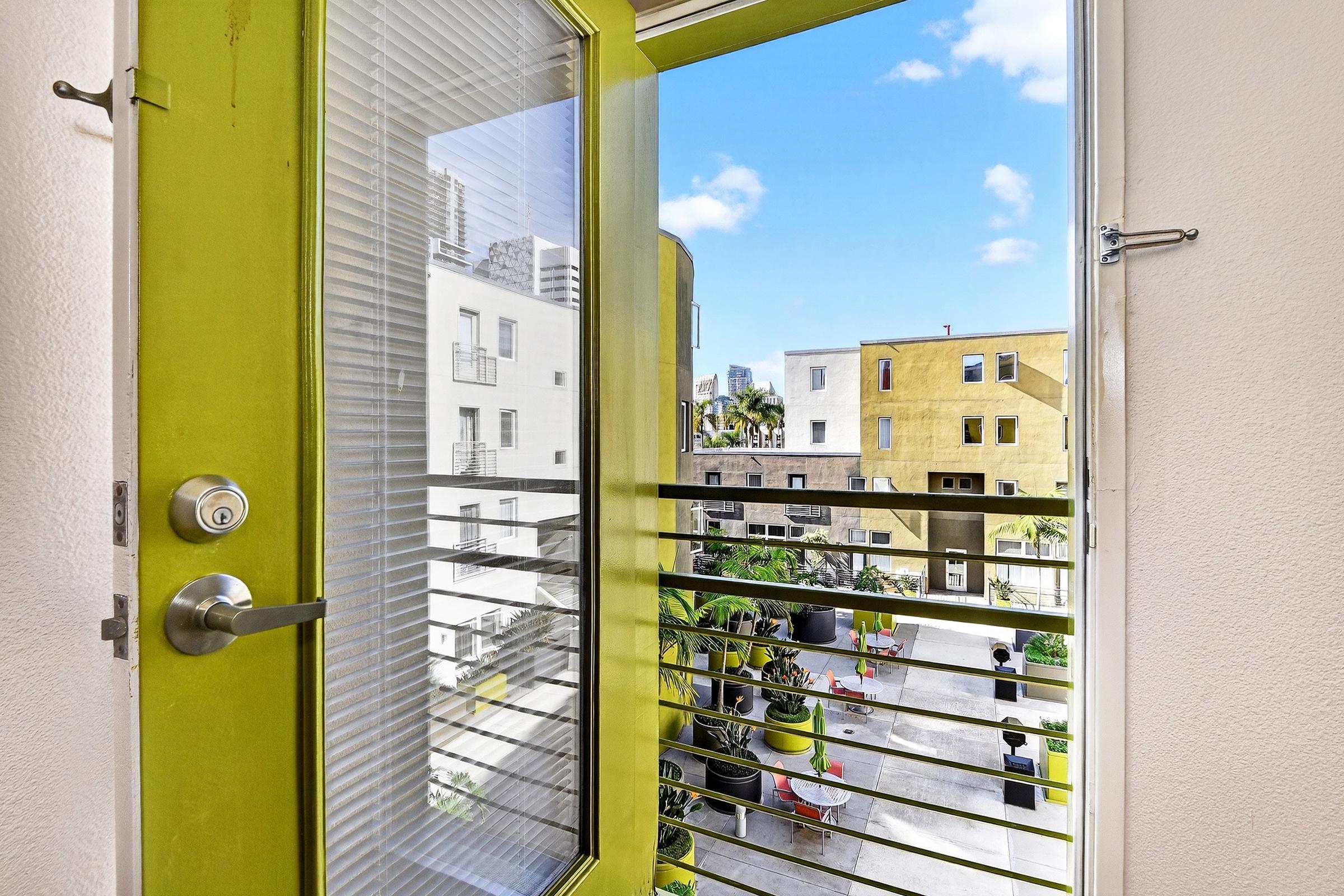 View from an open balcony door showcasing a sunny courtyard with greenery and modern buildings in the background. The door frame is painted in vibrant green, contrasting with the light walls and the blue sky.