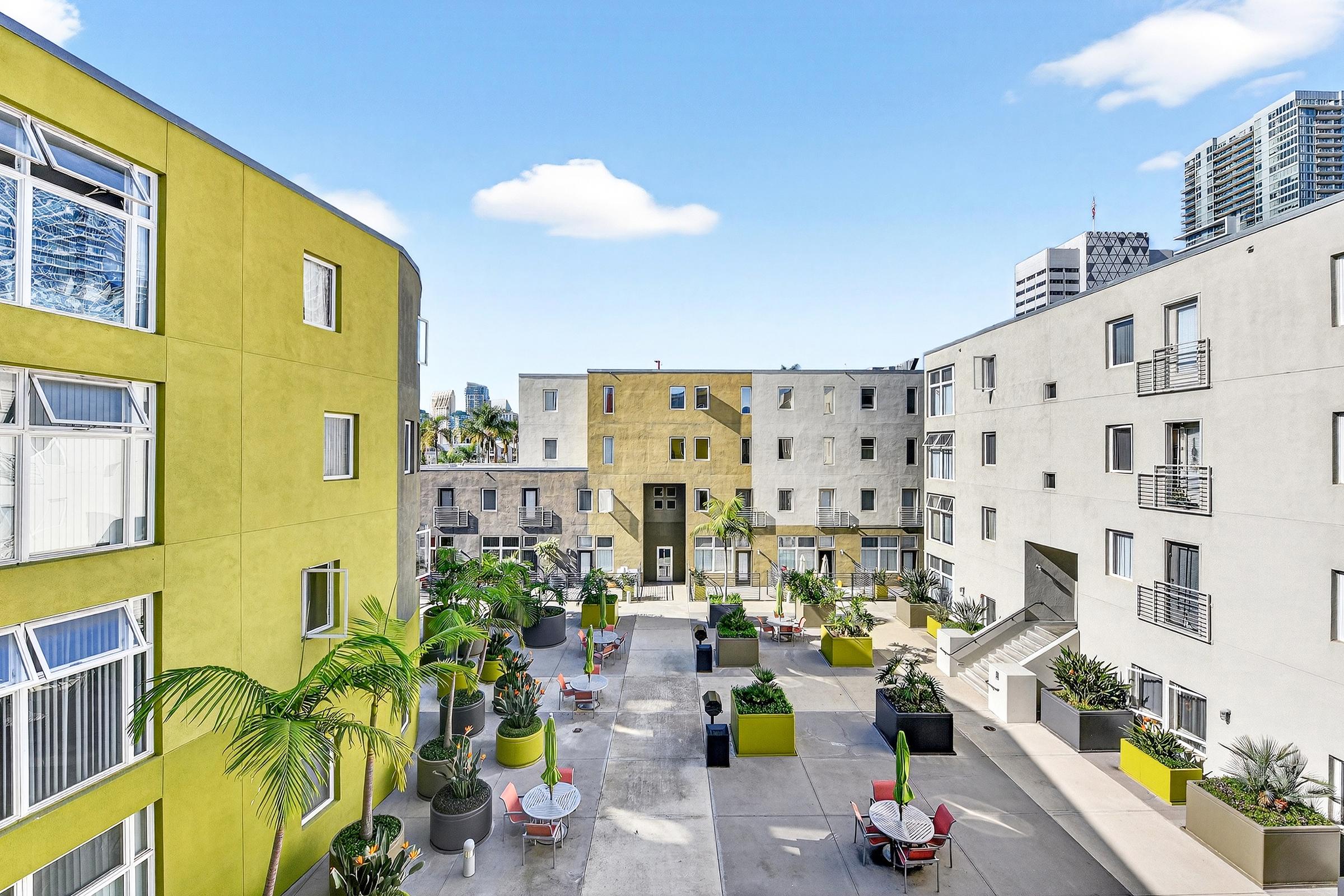A vibrant urban courtyard surrounded by modern apartment buildings. The buildings feature a mix of yellow and gray facades. Lush green plants and colorful outdoor furniture create a welcoming space for relaxation. Clear blue skies above enhance the bright atmosphere.