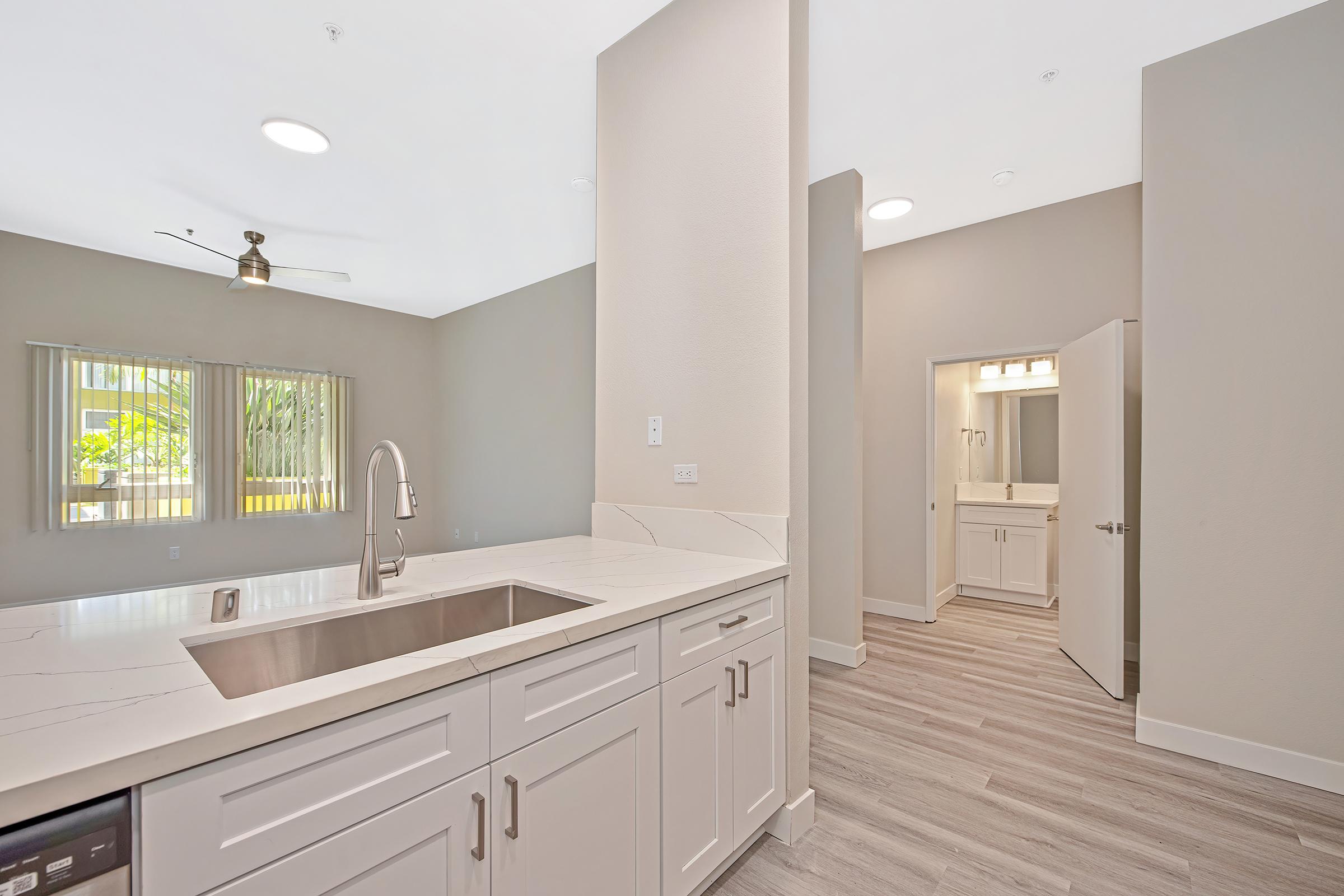 Modern kitchen and dining area with a large sink, light countertops, and under-cabinet lighting. The room features light wood flooring and beige walls. A doorway leads to a bathroom with bright lighting, and large windows provide natural light, showcasing greenery outside.