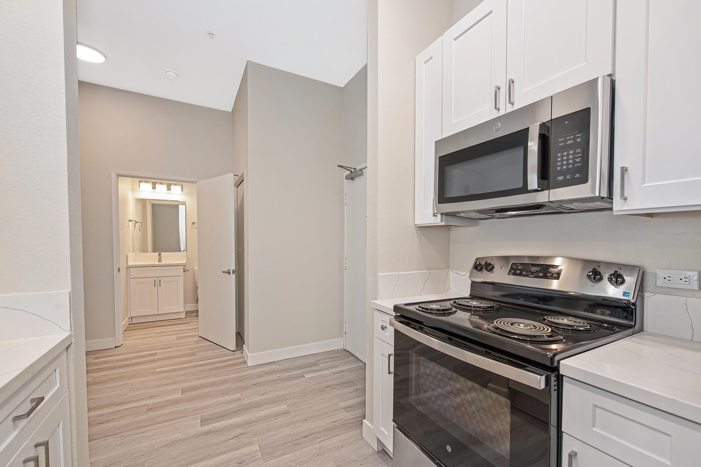 Modern kitchen with white cabinets, stainless steel appliances including a microwave and oven, and light-colored flooring. A doorway leads to a bathroom with bright lighting, and the walls are painted in a soft neutral tone. The space is well-lit and features a clean, contemporary design.