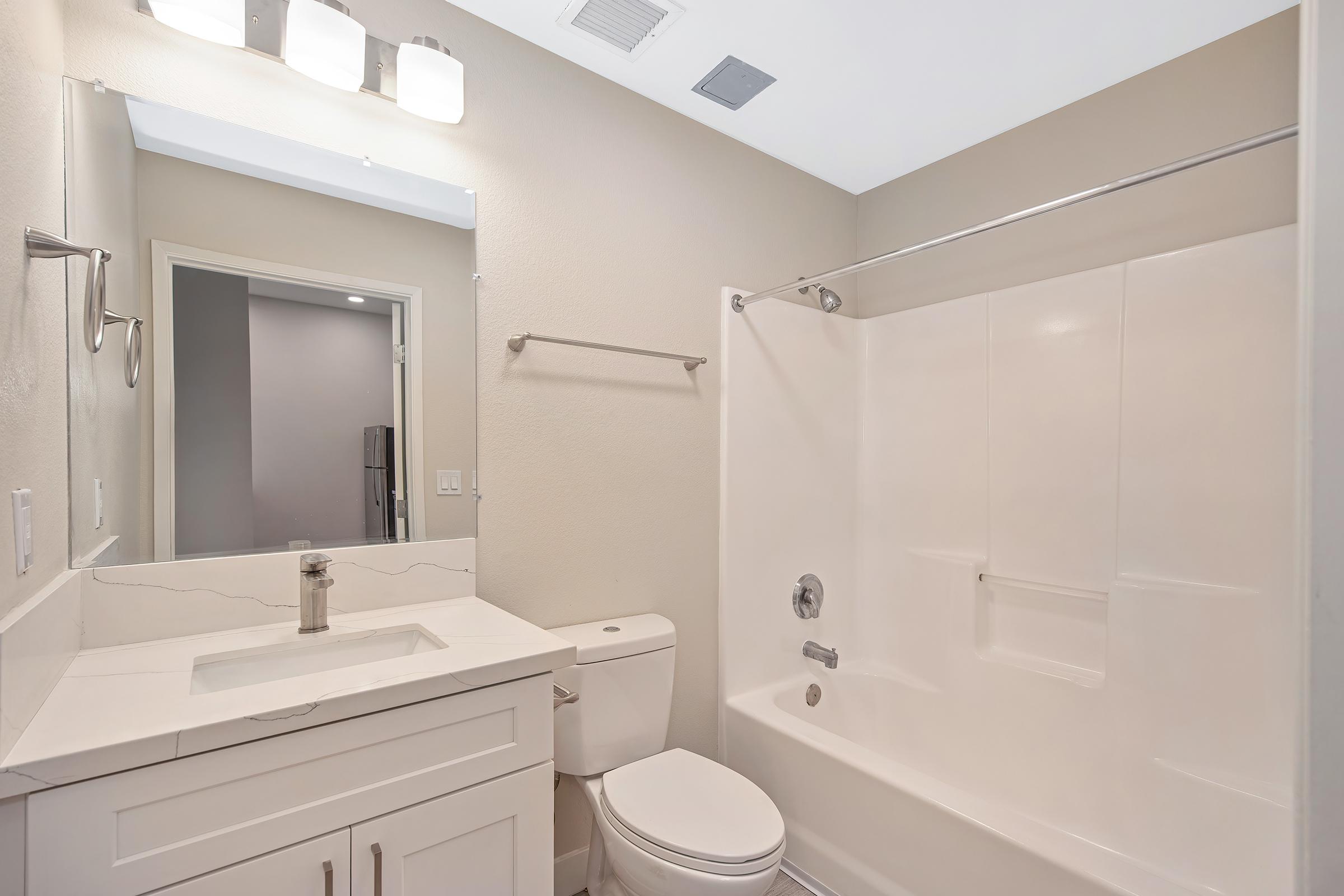 A modern bathroom featuring a white bathtub with a shower, a sleek sink with a faucet on a marble countertop, a large mirror above the sink, and a toilet. The walls are painted in a neutral tone, and there are light fixtures above the mirror providing ample lighting.