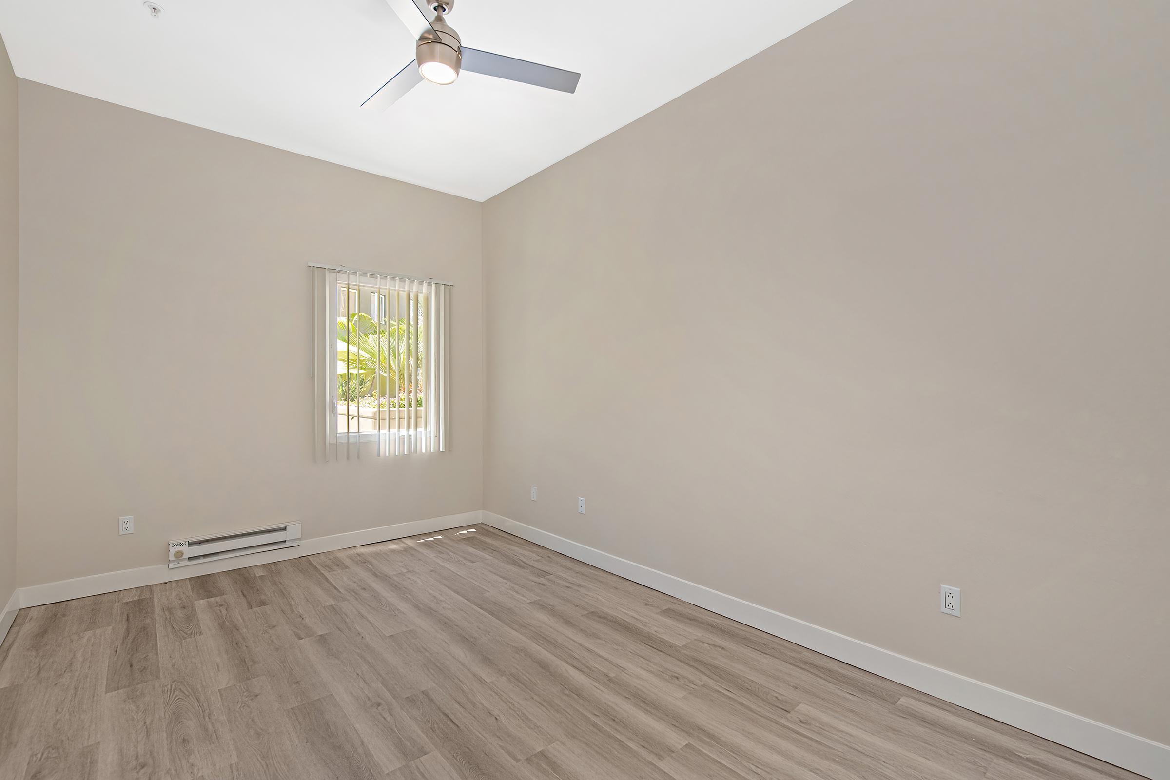 Empty room with light-colored walls and wood-look flooring. A ceiling fan is mounted in the center, and a window with vertical blinds allows natural light to enter. The room appears spacious, with minimal furnishings and a simple, uncluttered design.