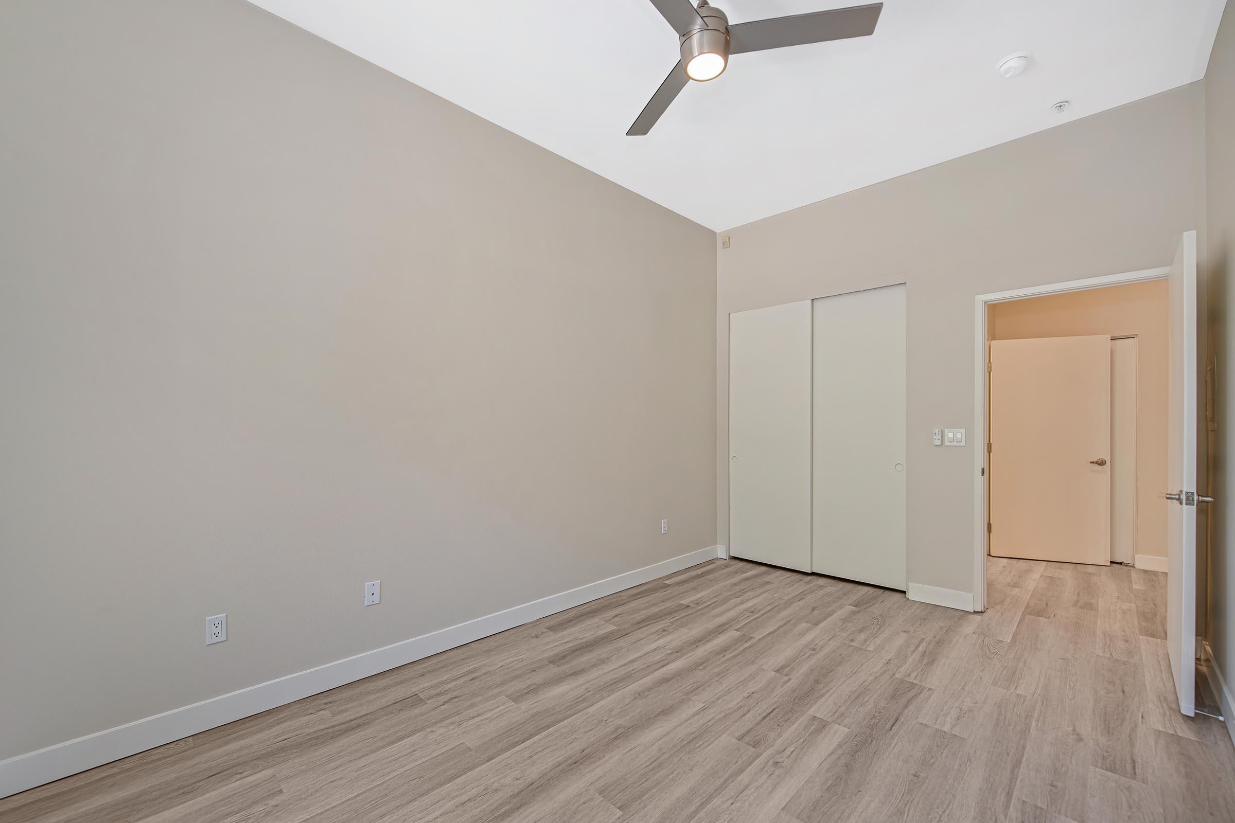 A spacious, empty room with light-colored walls and wood-like flooring. There is a modern ceiling fan and a closet with bi-fold doors on one side. A doorway leads to another room, and there is ample natural light coming in from the ceiling. The room is clean and ready for furnishings.
