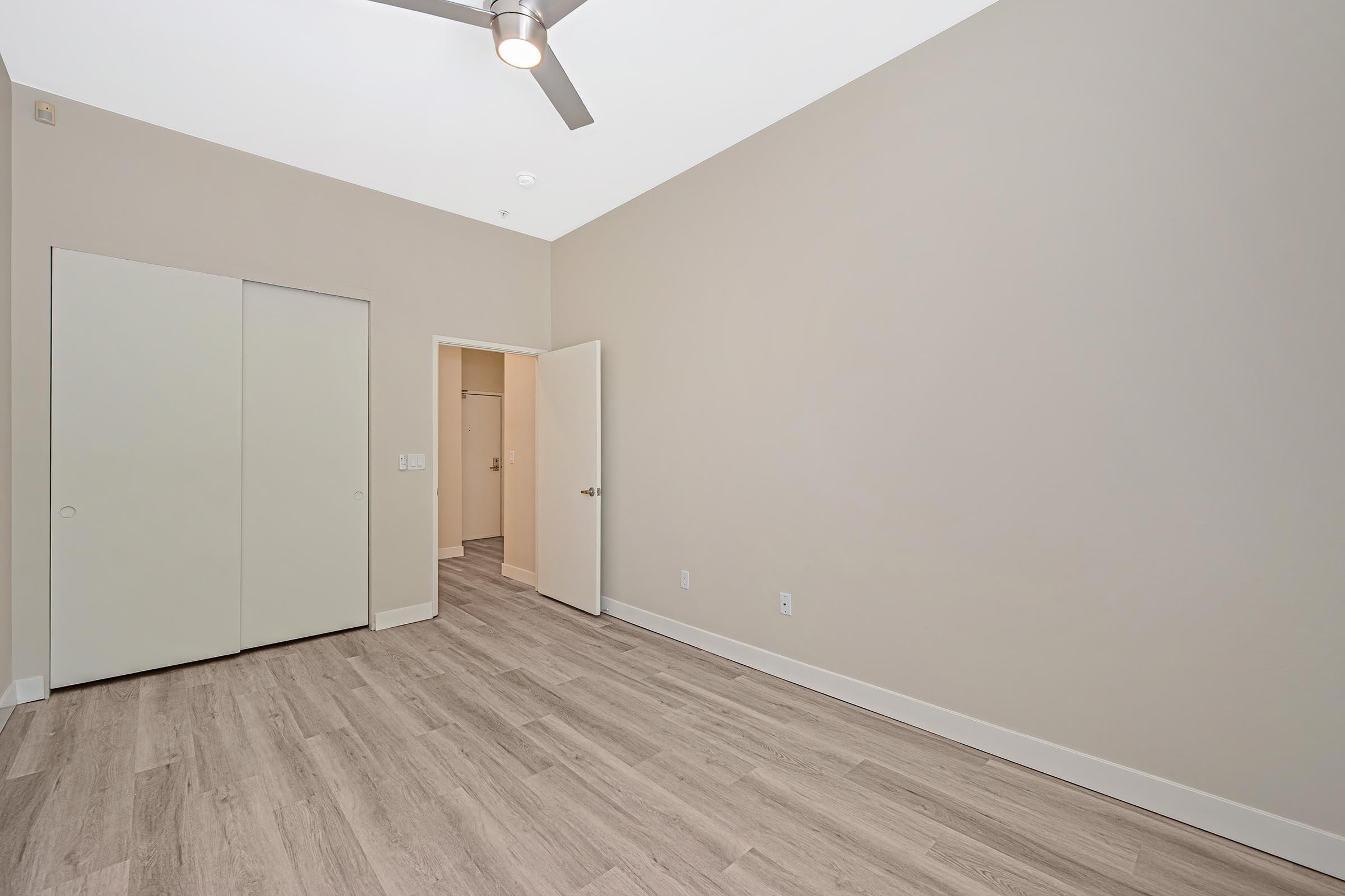 Interior view of an empty room featuring light beige walls and a wooden floor. A ceiling fan is visible, and there is a closed double door closet on the left. An open door leads to another room, hinting at a spacious layout. Natural light enhances the bright and airy feel of the space.
