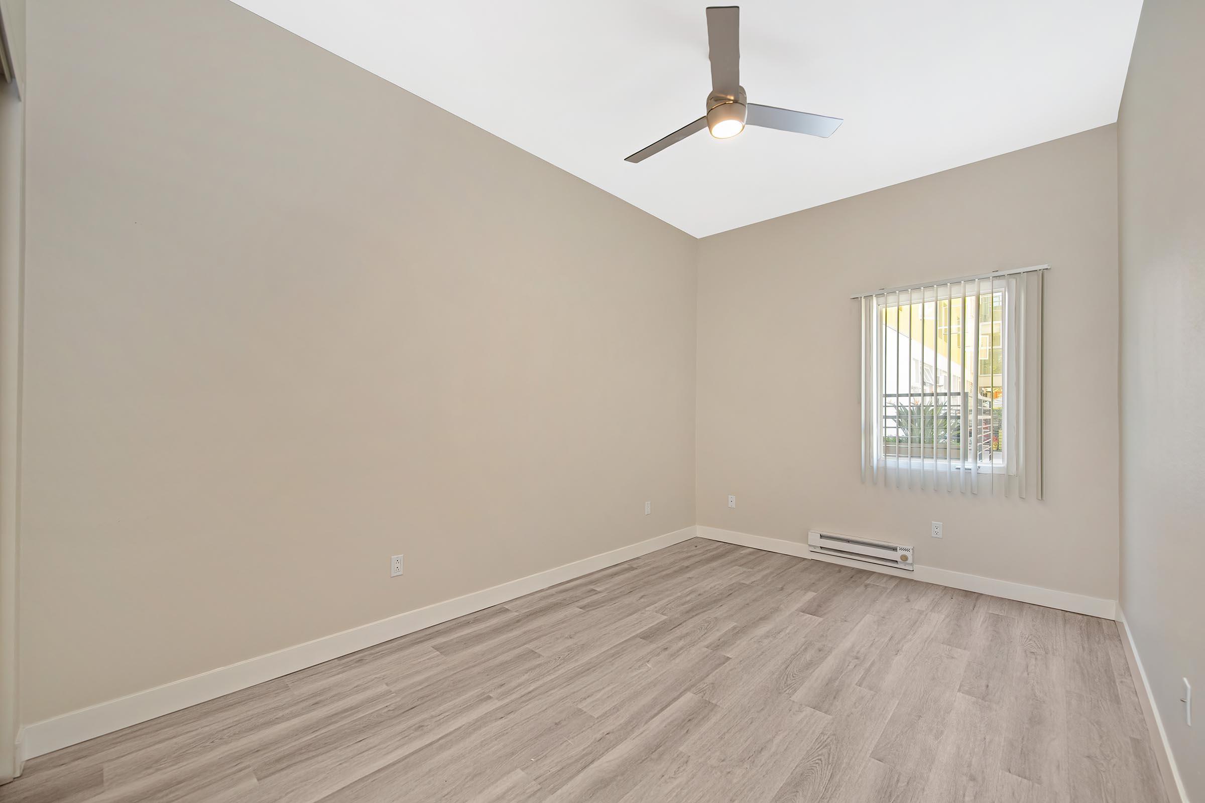 A spacious, empty room with light-colored walls and a ceiling fan. The floor is covered with light wood-like laminate. There is a window with vertical blinds allowing natural light to enter the room, and a baseboard heater visible near the floor. The overall atmosphere is bright and airy.