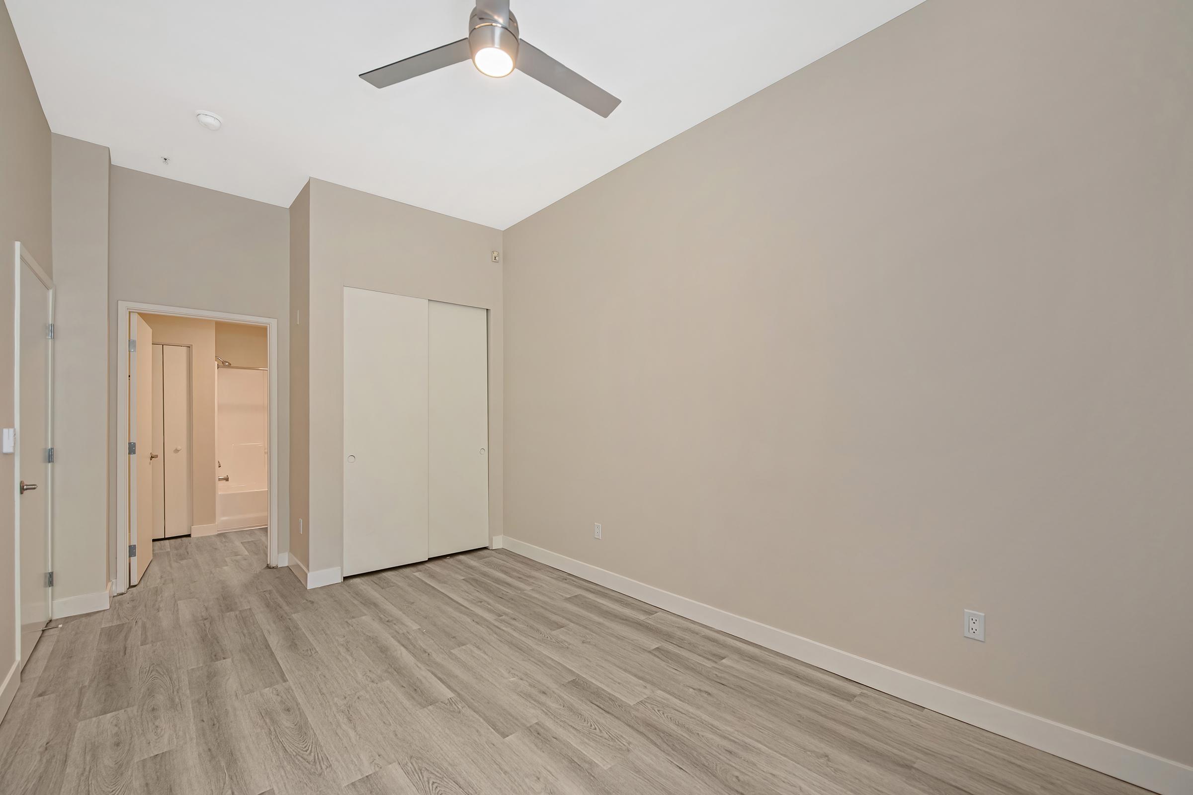 A neutral-colored room with a ceiling fan and light wood flooring. There are two closed closet doors on one wall, and an open doorway leading to a bathroom with a shower visible in the background. The walls are plain and unadorned, creating a spacious feel.