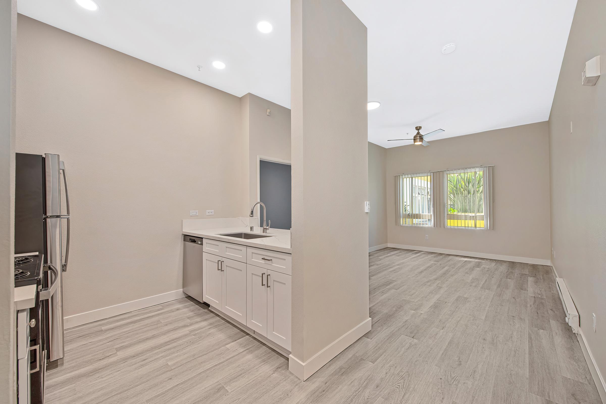 Modern kitchen and living area with light-colored walls and wood-style flooring. The kitchen features stainless steel appliances, white cabinets, and a large window providing natural light. The living space has a ceiling fan and large windows overlooking greenery.