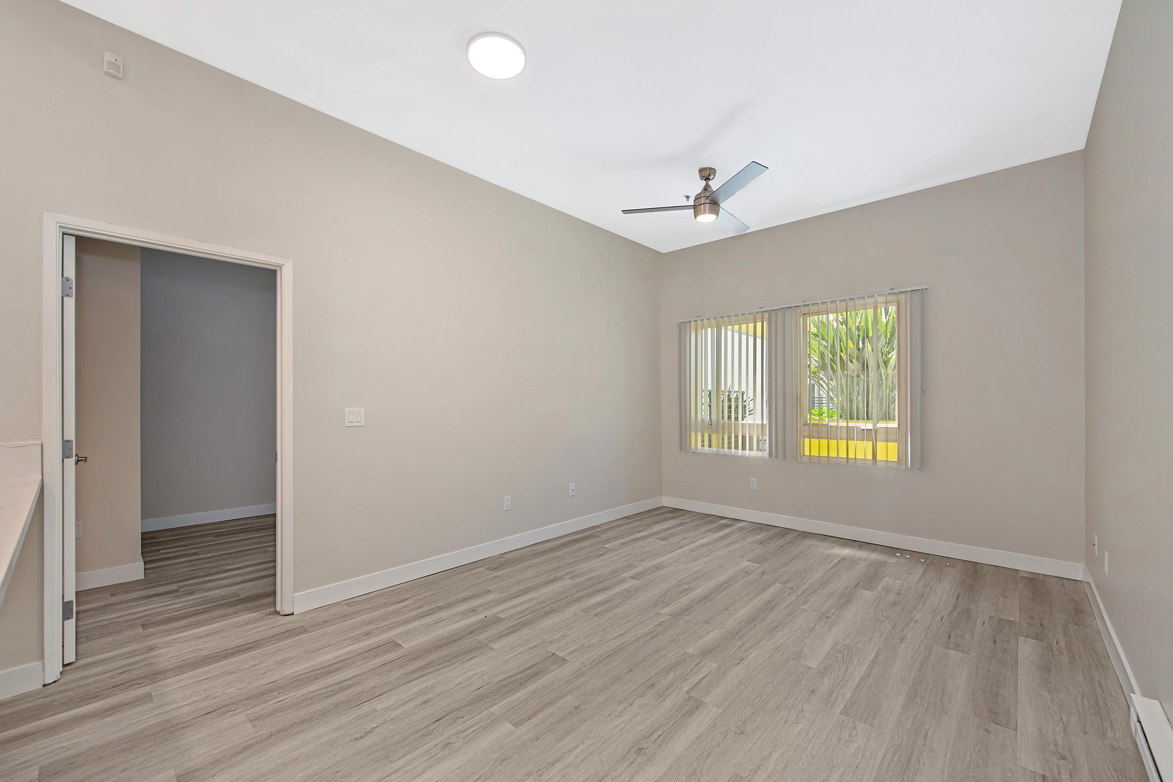A spacious, empty room with light gray walls and wooden flooring. A ceiling fan is visible, and there's a window with blinds allowing natural light. A doorway leads to another area, enhancing the openness of the space. The room is well-lit and has a modern design.