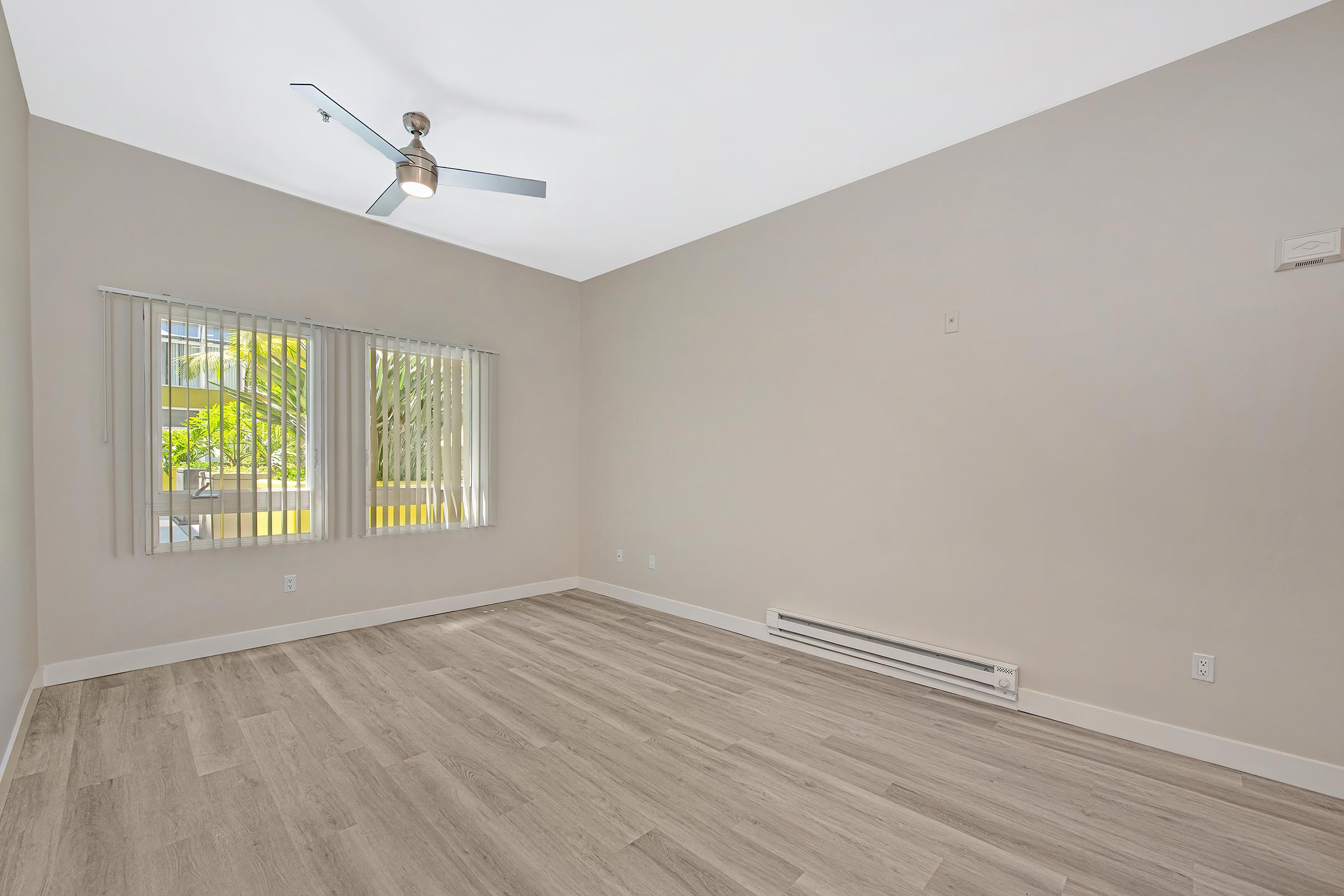 A spacious, empty room with light-colored walls and wooden flooring. It features a ceiling fan and large windows with blinds, allowing natural light to enter. A baseboard heater is visible along one wall, highlighting the room's simplicity and openness.