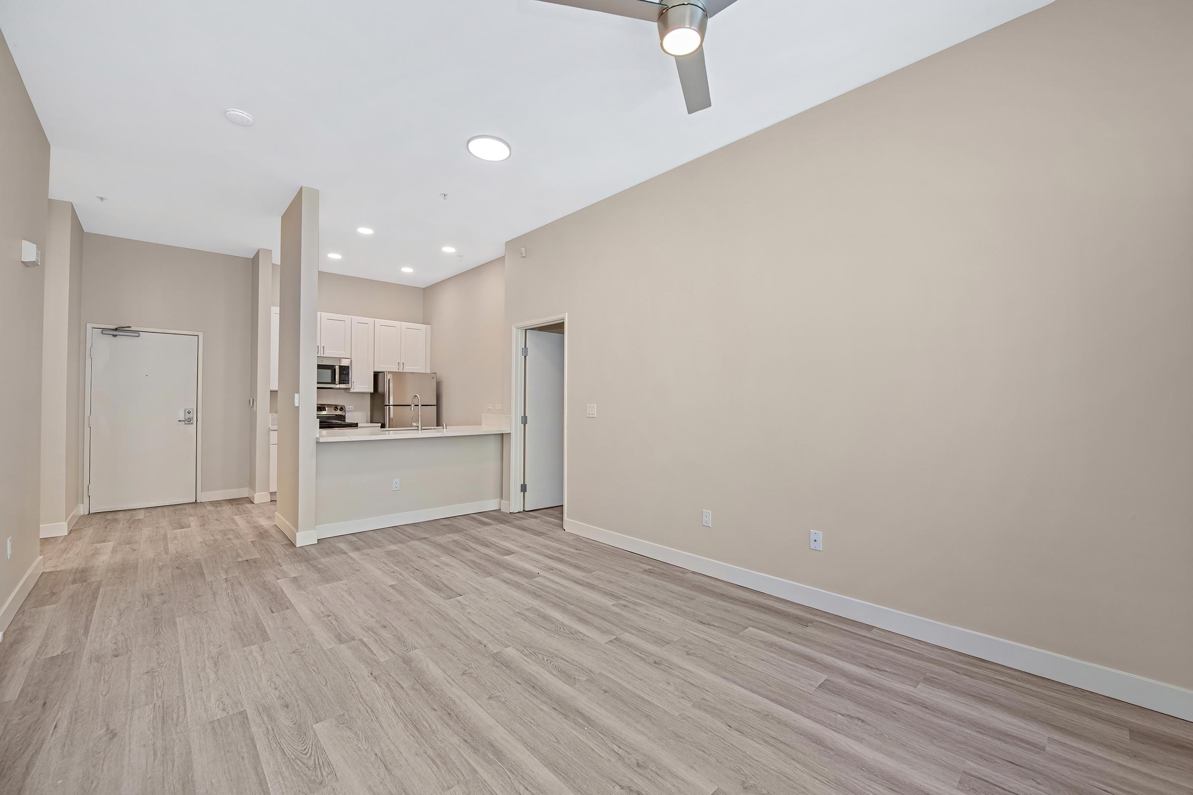 A spacious, modern living area with pale walls and wood-like flooring. The room features a ceiling fan, an open kitchen with light cabinetry and stainless steel appliances, and a door leading to the outside. The space is well-lit with natural light coming from unseen windows.