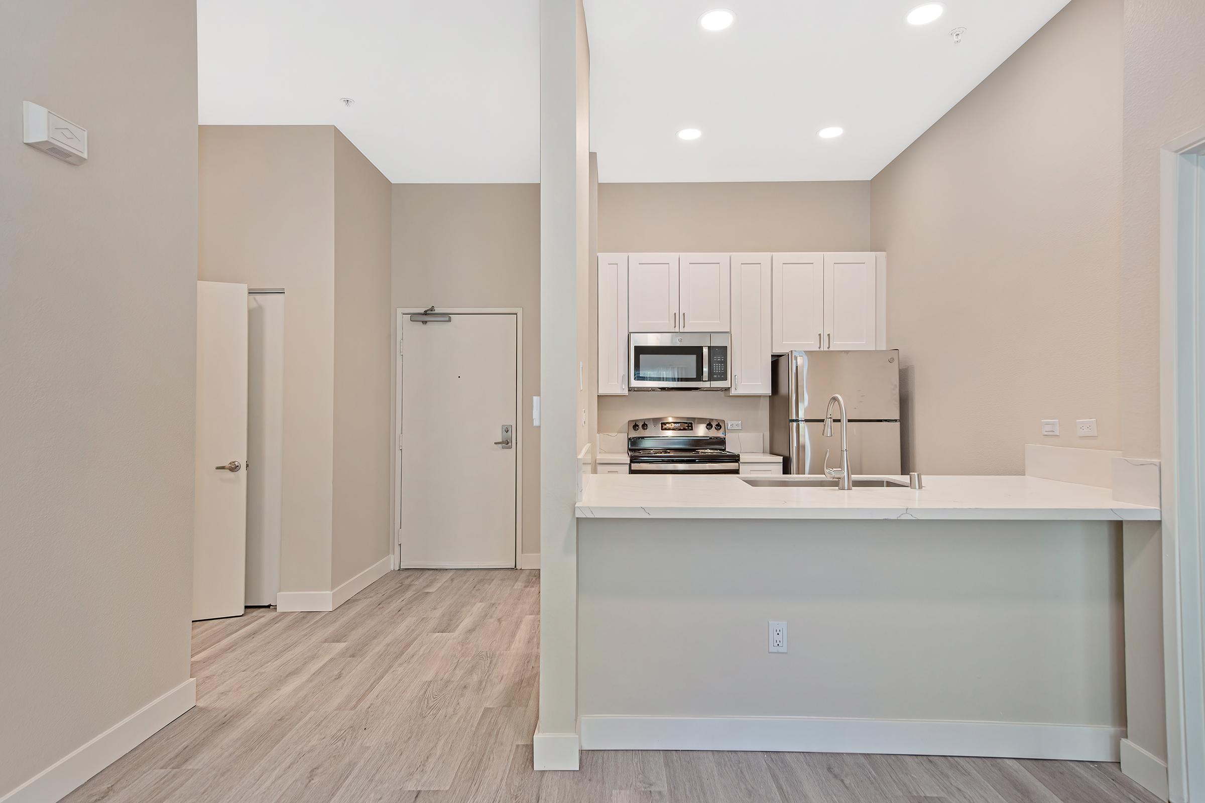 Modern kitchen and living area with light-colored walls and wooden flooring. The kitchen features white cabinetry, stainless steel appliances, and a breakfast bar. A door leads to a hallway, and natural light fills the space from recessed lighting above.