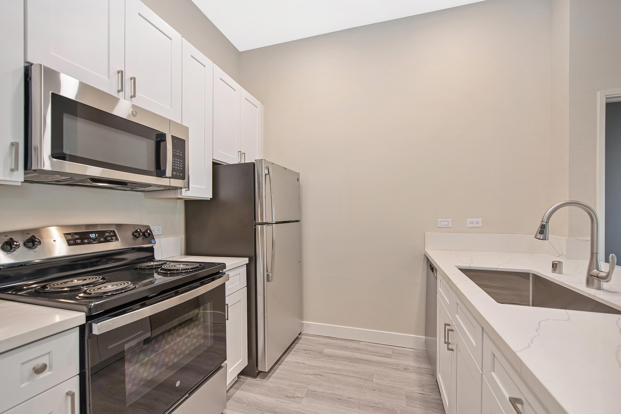 Modern kitchen featuring stainless steel appliances including a microwave, range, and refrigerator. The cabinetry is white with a sleek design, and the countertop is a light-colored stone. The floor is light gray, contributing to a bright and clean aesthetic.