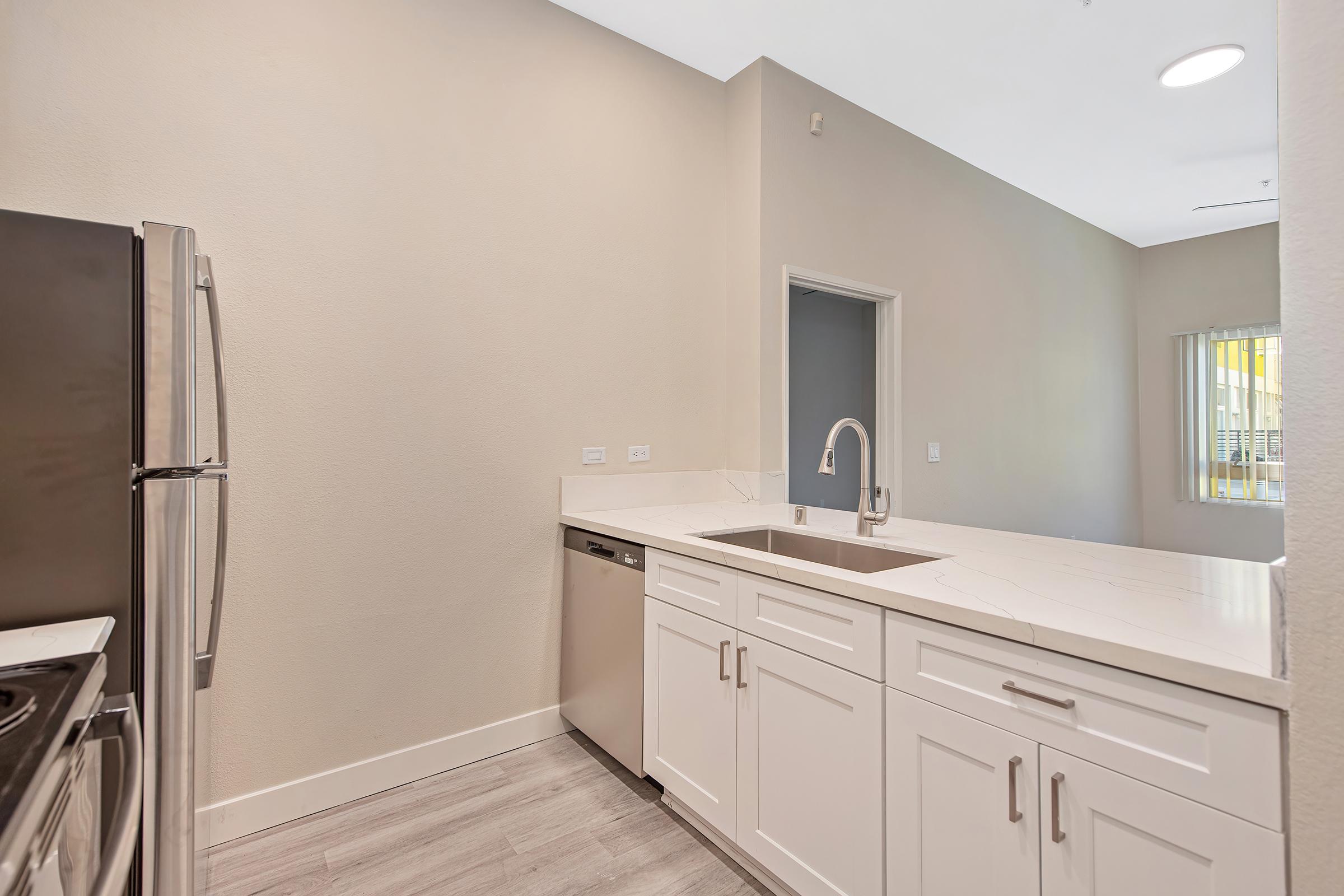 Modern kitchen featuring stainless steel appliances, including a refrigerator and dishwasher, light-colored cabinets, and a quartz countertop. The design includes an open layout with a neutral wall color and ample natural light from a nearby window.