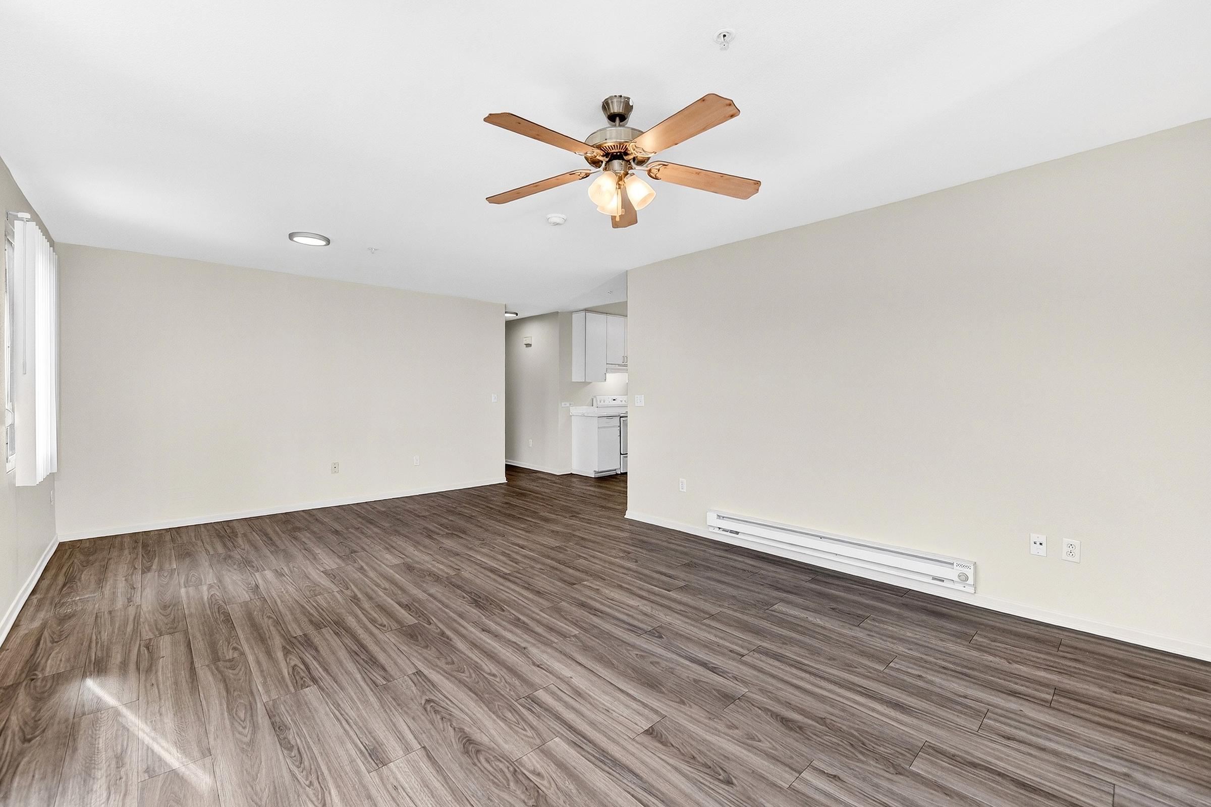Spacious living room with light brown laminate flooring, a ceiling fan with wooden blades, and neutral-colored walls. Natural light streams through a window, creating a bright atmosphere. In the background, a kitchen area is partially visible, featuring white cabinetry.
