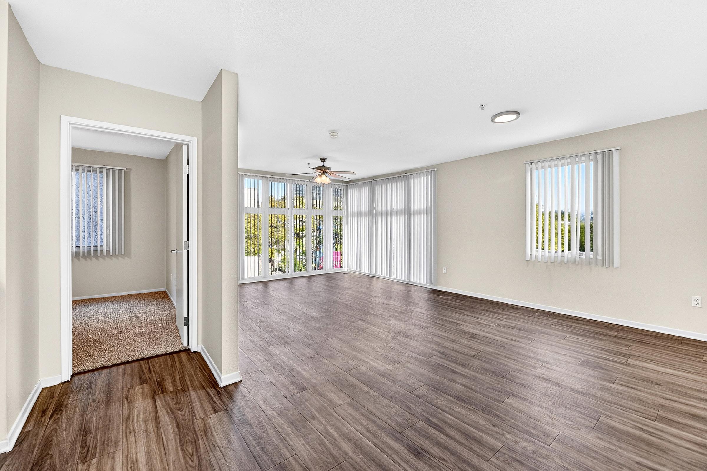 Spacious living area with wood-style flooring, large windows allowing natural light, and ceiling fan. An adjacent room features carpet flooring and a doorway leading outside. Vertical blinds on the windows enhance the airy atmosphere. Modern, neutral color scheme throughout.