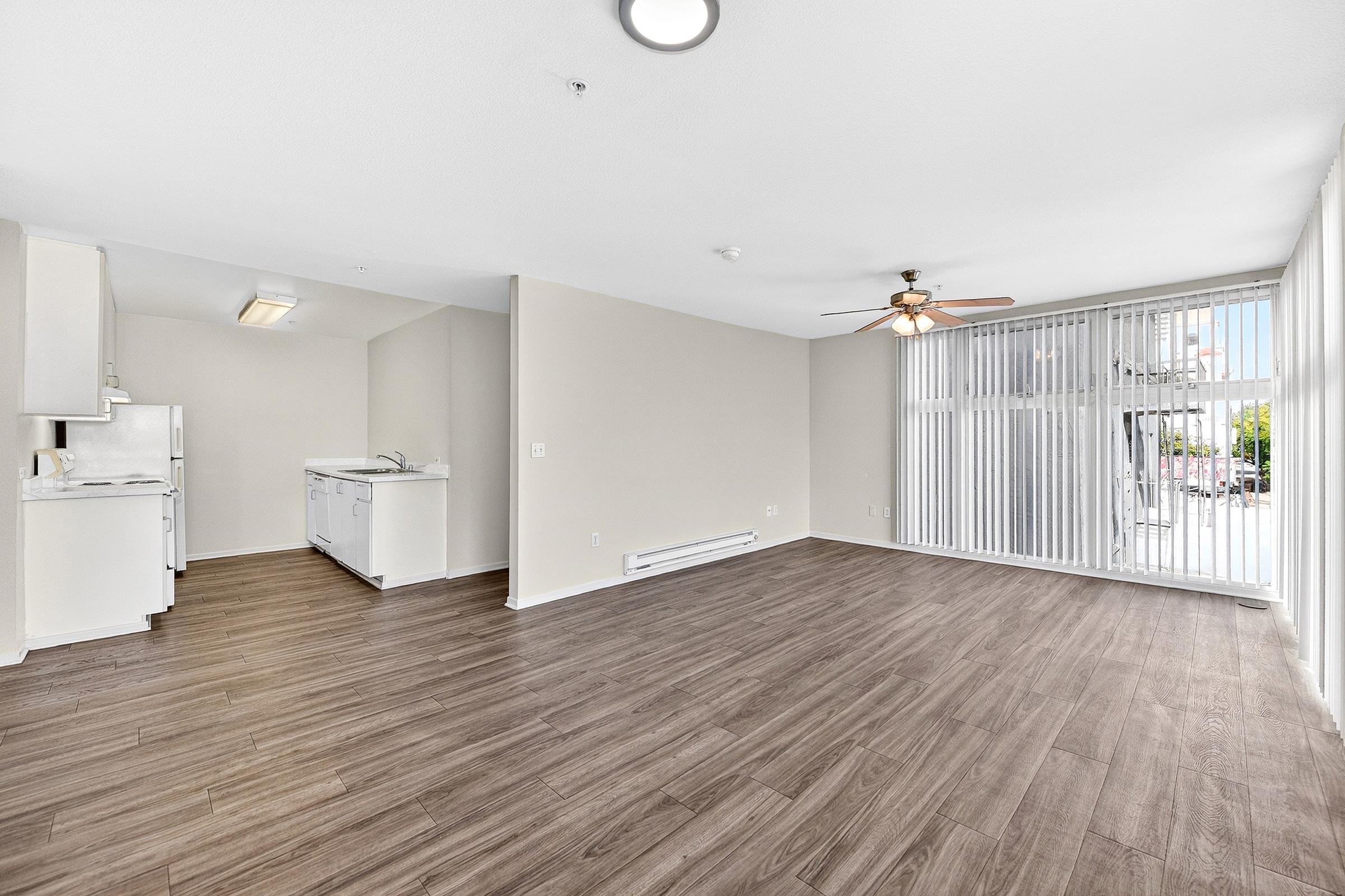 Spacious living area with wood laminate flooring, featuring a ceiling fan and large windows with vertical blinds. Open kitchen visible on the left with white appliances and cabinetry. Well-lit and neutral-colored walls, creating a bright and airy atmosphere.