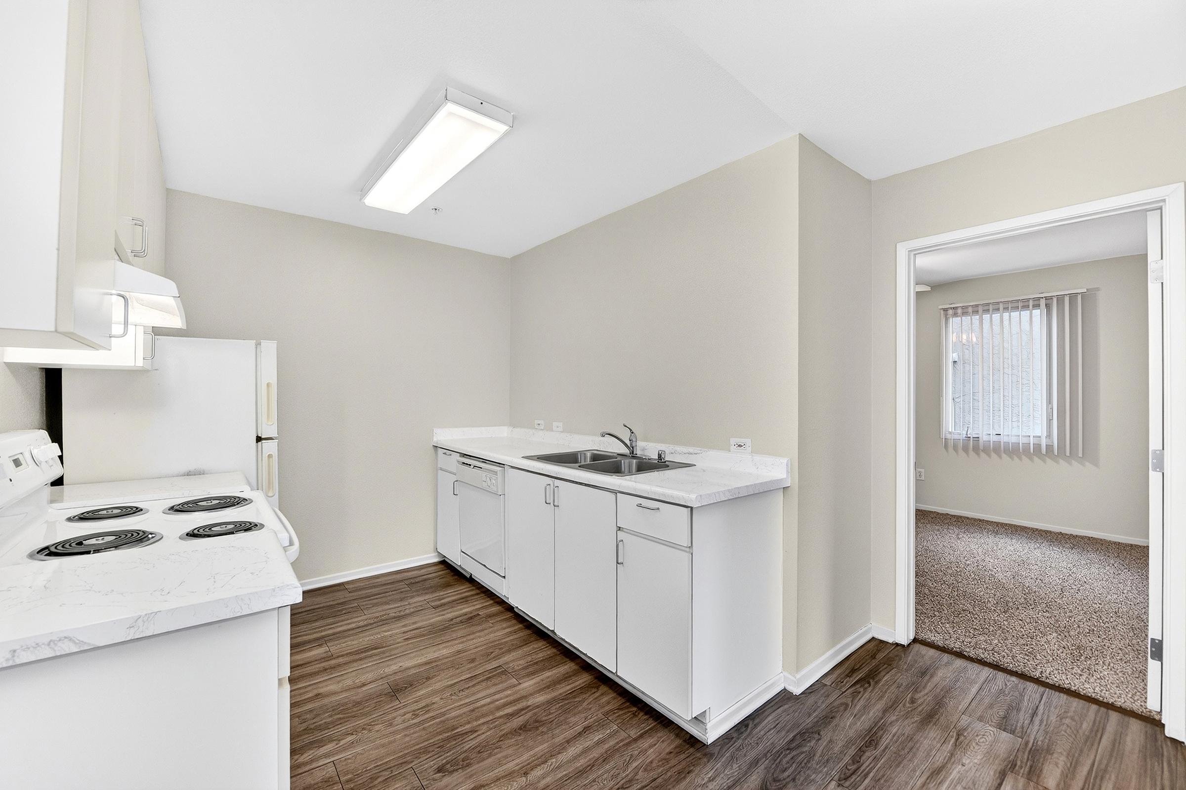 A modern kitchen featuring white cabinetry, a double sink, and a countertop. The kitchen includes an electric stove and refrigerator. Light-colored walls and a large window provide natural light. An adjacent doorway leads to a carpeted living area with vertical blinds.