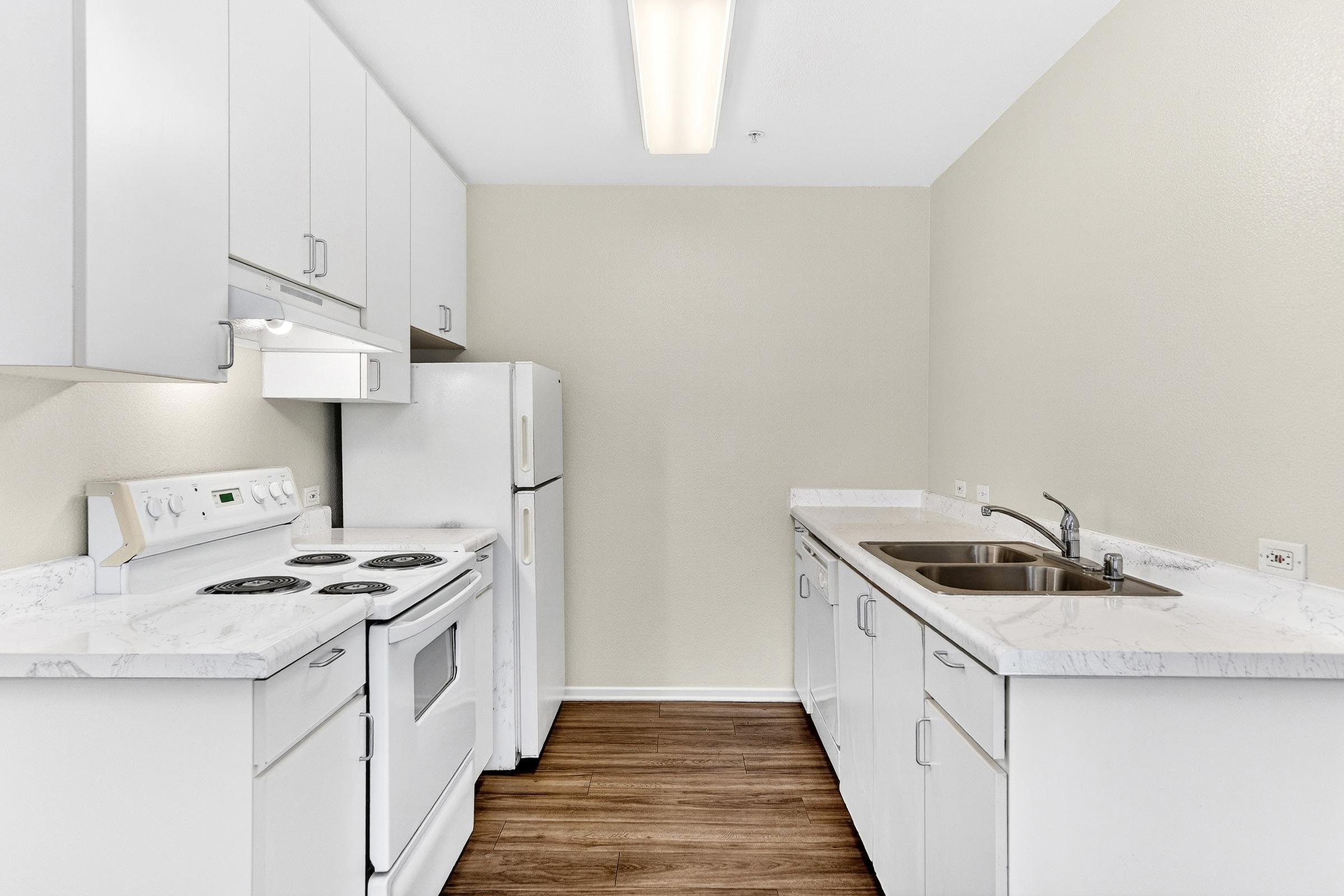 A clean, modern kitchen featuring white cabinets, a double sink, an electric stove, and a refrigerator. The countertops have a marble-like finish, and the space is illuminated by a ceiling light, creating a bright atmosphere. Wood-like flooring adds warmth to the design.