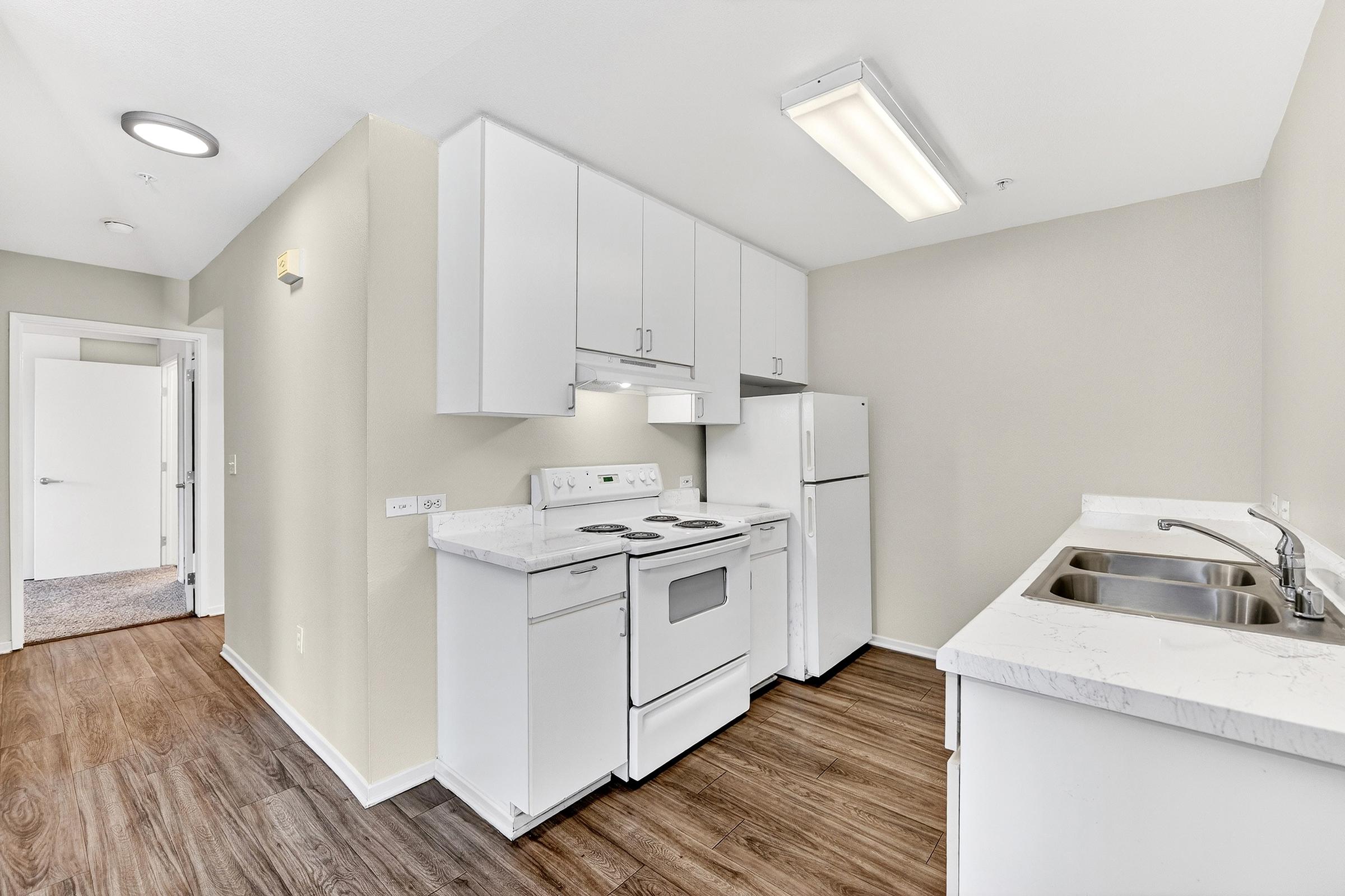 A modern kitchen featuring white cabinets, a white stove, oven, and refrigerator. The countertop is light-colored with a double sink. The walls are painted a neutral shade, and there is a ceiling light. A doorway in the background leads to an adjoining room. The flooring is wood-like laminate.