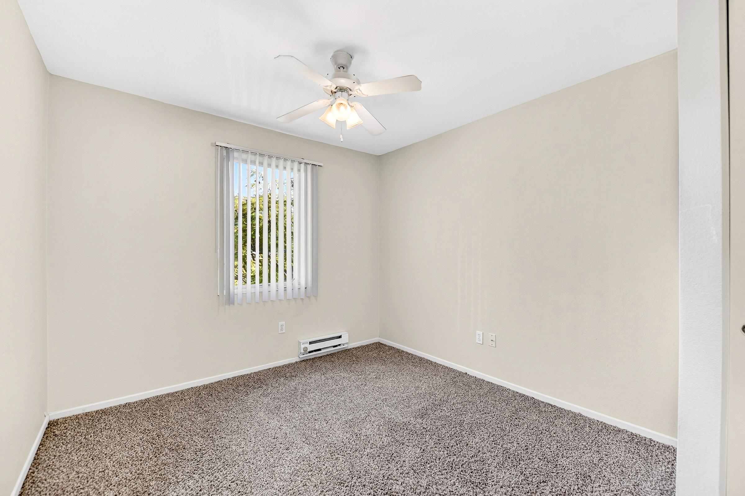 A simple, empty room featuring light beige walls and carpet, equipped with a ceiling fan. A window with vertical blinds allows natural light, and there is a baseboard heater. The space is uncluttered and ready for furnishing.