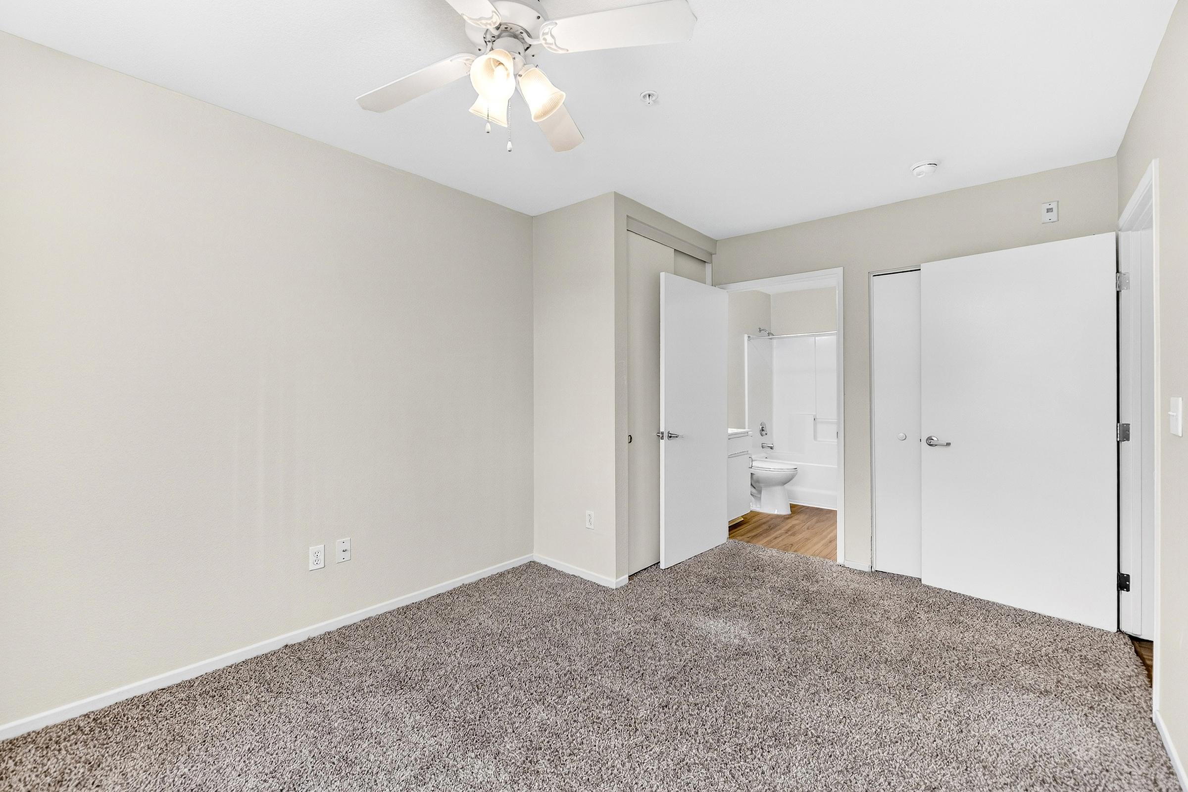 An empty room with light-colored walls and a ceiling fan. The floor is covered in carpet. Two closed doors lead to an adjacent bathroom and closet. Natural light enters from a window, illuminating the space. The room is clean and unfurnished, ideal for customization.