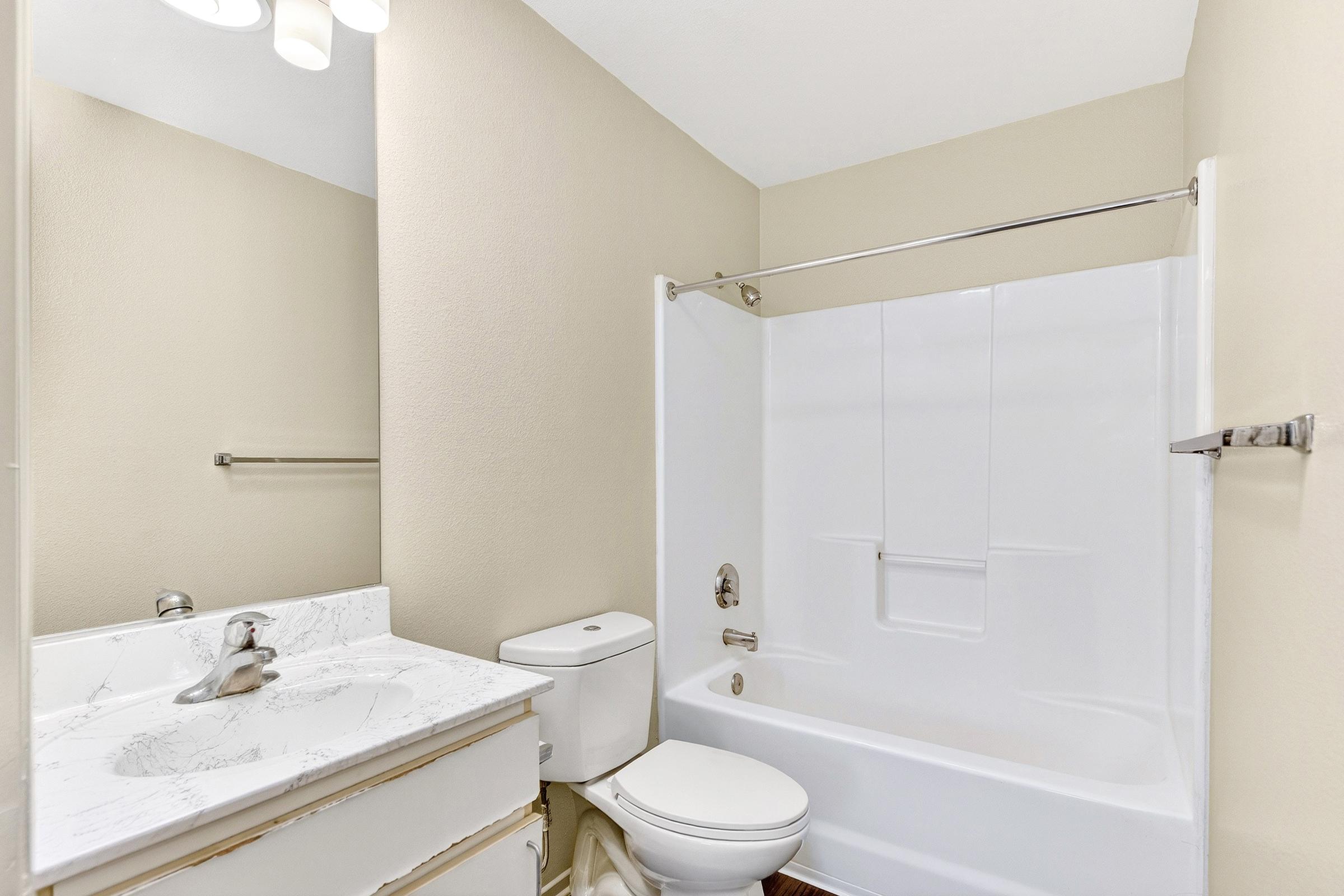 A clean, well-lit bathroom featuring a white bathtub with a shower, a modern sink with a marble countertop, a toilet, and a large mirror. The walls are painted in a light beige color, complemented by neutral tones in the fixtures and flooring.