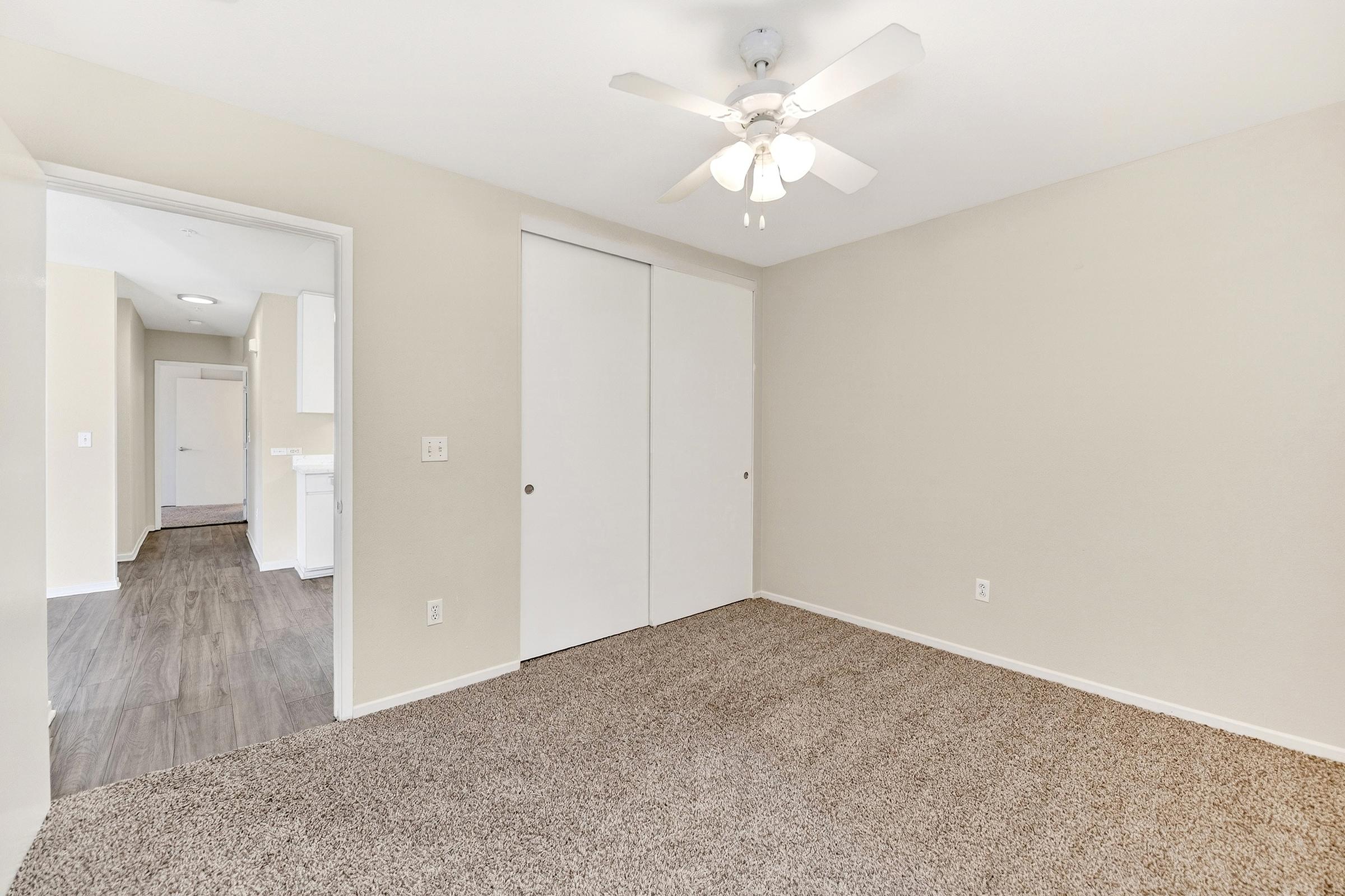 A spacious, empty room featuring light-colored walls and a ceiling fan. The floor is covered in plush carpet. In the background, a doorway leads to another area, highlighting a hallway and part of a kitchen with white cabinetry. Natural light brightens the space, creating an inviting atmosphere.