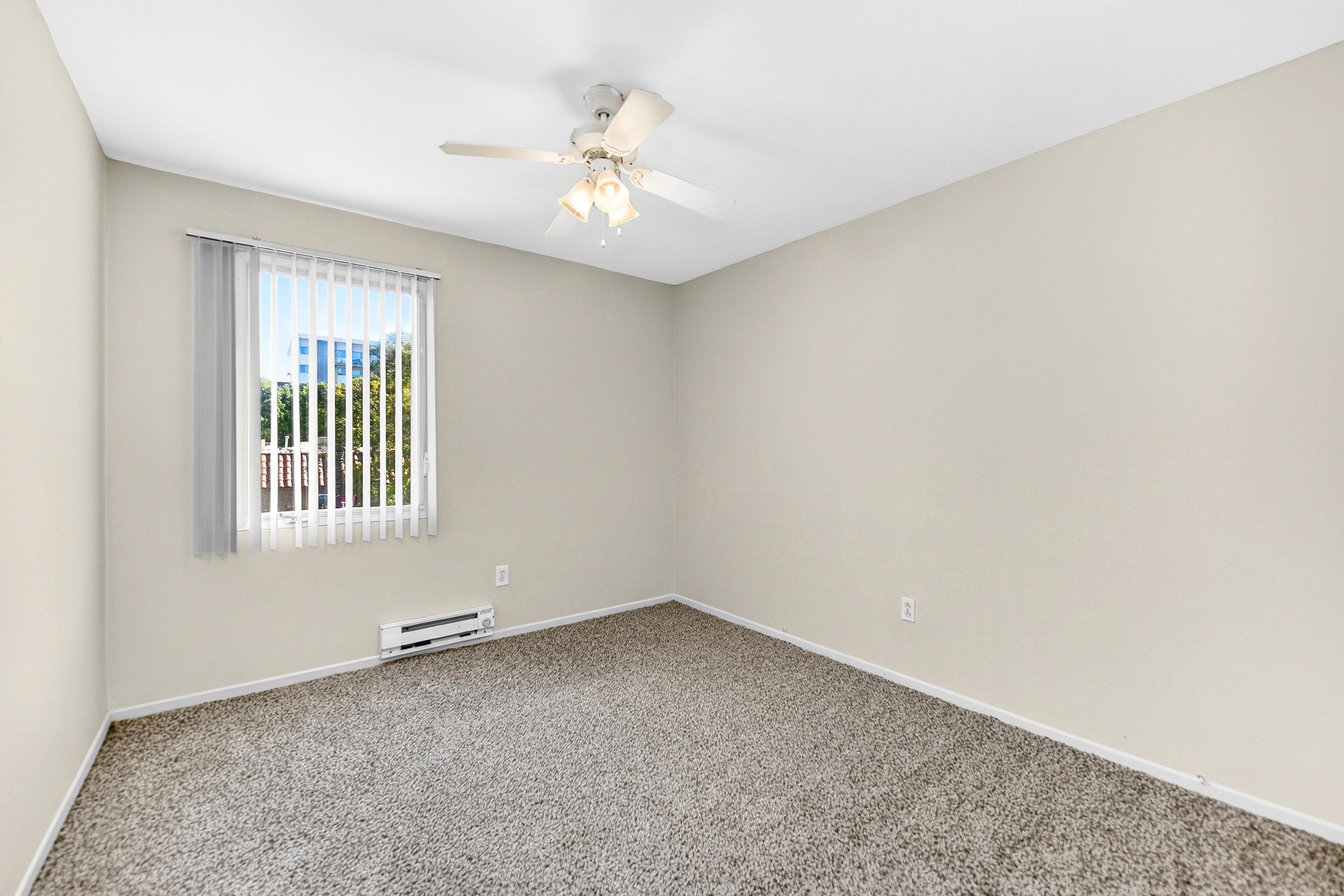 Empty room with light beige walls and carpeted flooring. A ceiling fan with lights is mounted in the center. There is a window with vertical blinds, letting in natural light, and an air conditioning unit on the wall. The overall space appears clean and ready for furnishing.