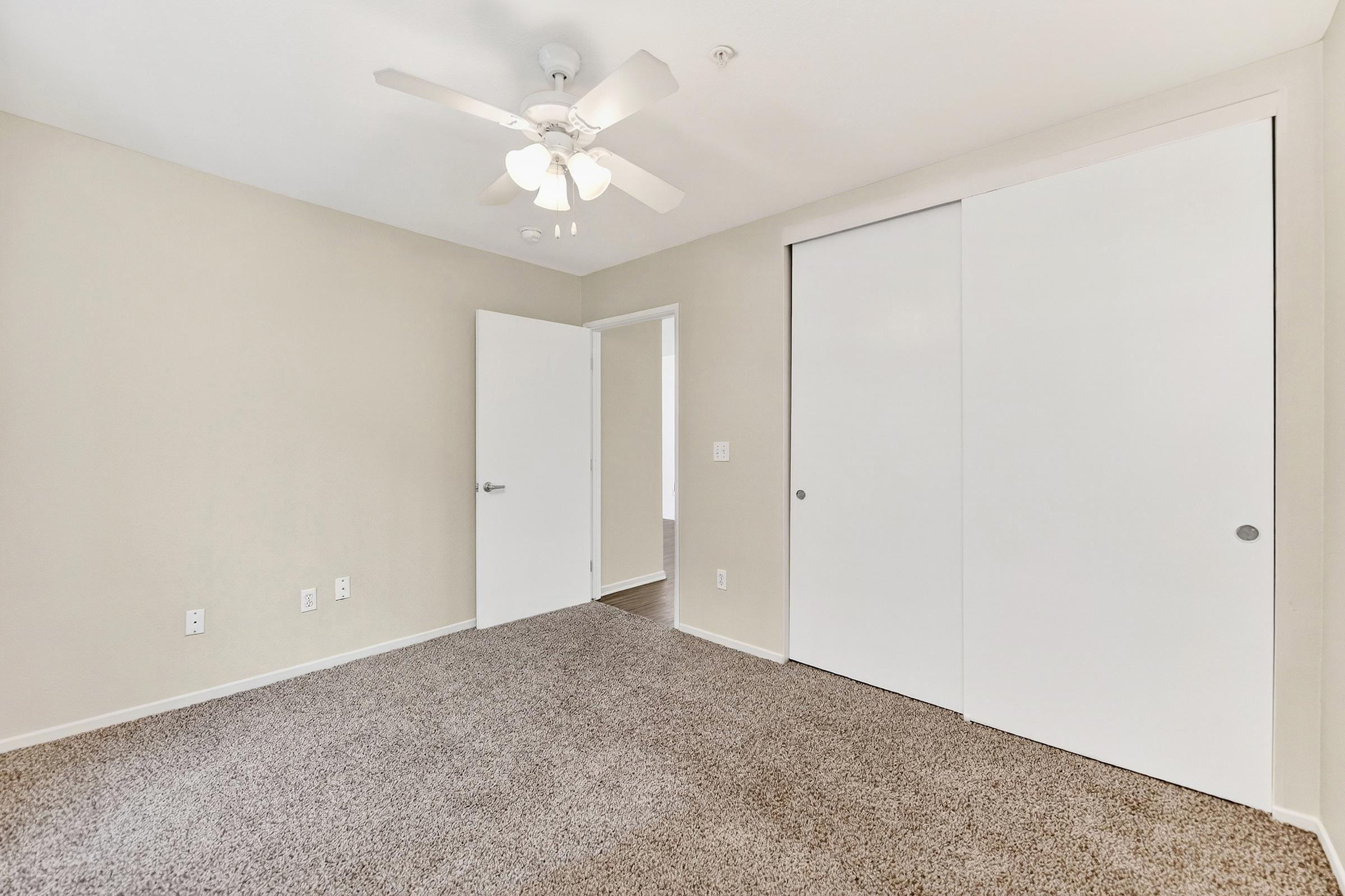 A small, empty room with beige walls and light brown carpet. A ceiling fan is mounted on the ceiling, and there are two white sliding closet doors on one wall. A door leads to another room, and the space is well-lit with natural light.