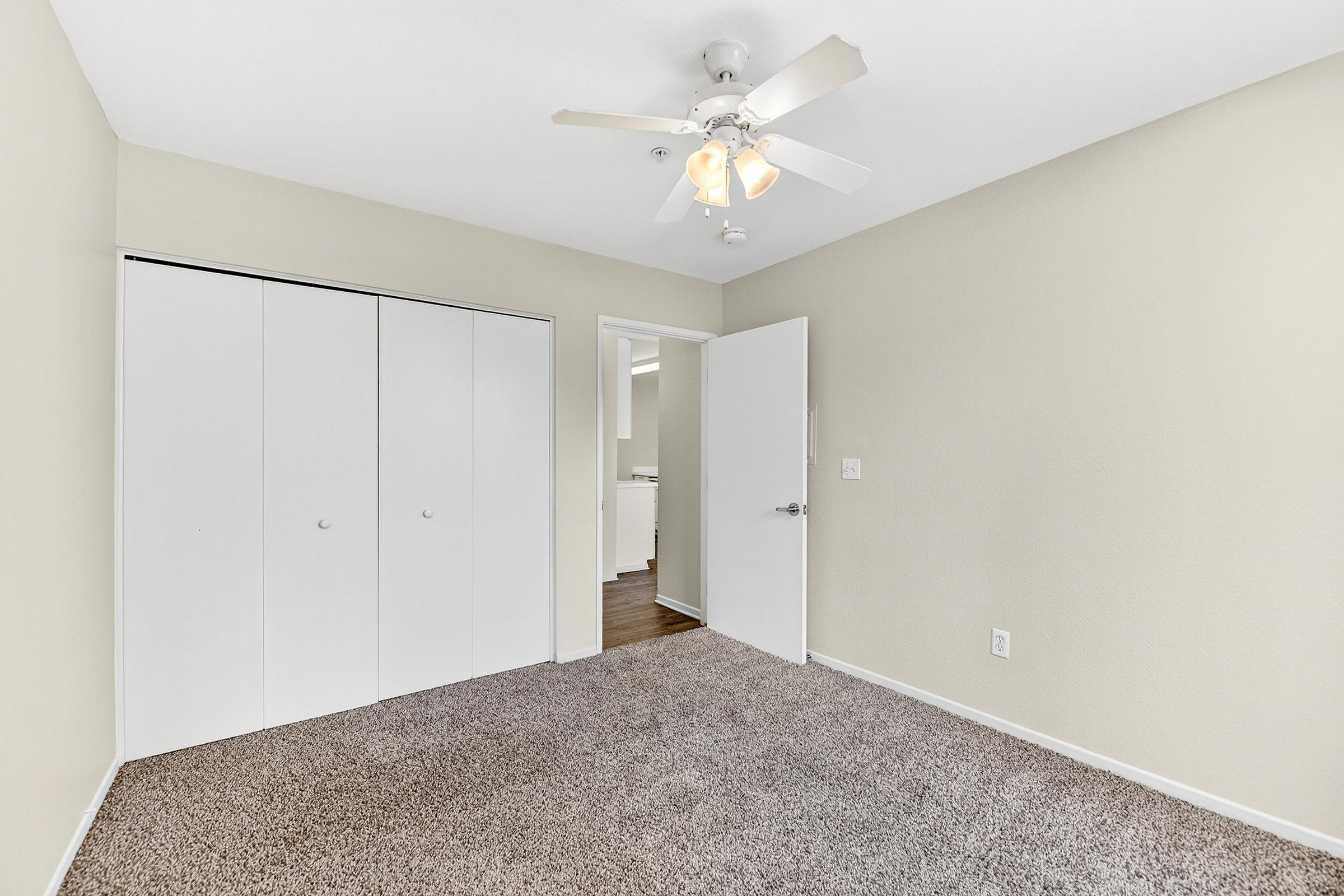 A well-lit bedroom featuring light beige walls, plush carpet, and a ceiling fan. There is a closet with sliding doors on the left side, and an open door leading to another room in the background. The space is tidy and vacant, ideal for new furniture or decor.